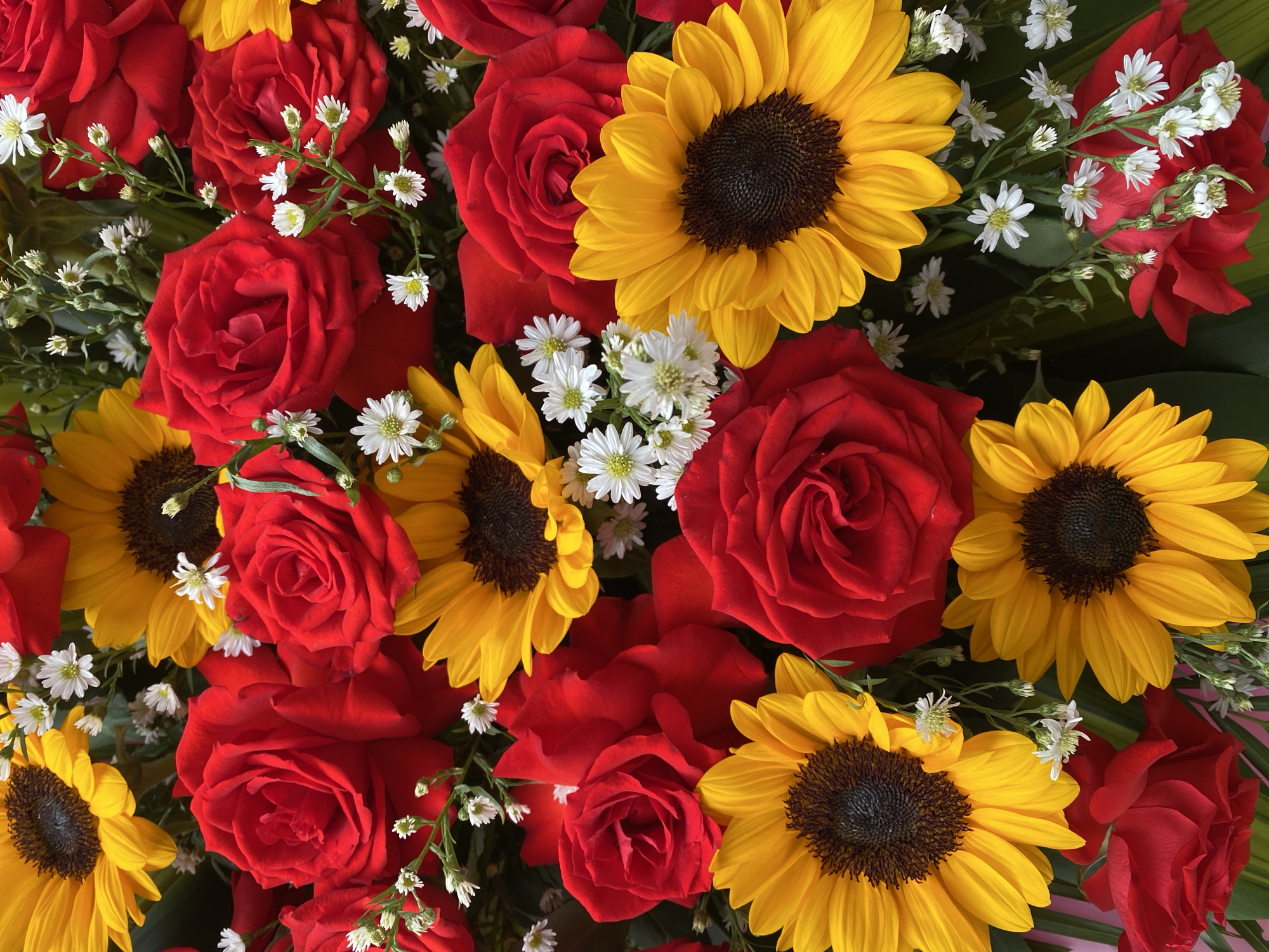 A close-up, overhead shot showcases a vibrant bouquet of red roses and yellow sunflowers, interspersed with delicate white filler flowers. The arrangement is densely packed, with the flowers filling the entire frame. The backdrop is a dark, leafy green, providing a rich contrast to the bright colors of the blooms. The lighting is soft and even, highlighting the textures and details of each petal. There are no people or actions depicted, just the static beauty of the floral arrangement. This image is likely from a flower shop or a special occasion in Đà Nẵng, Vietnam, given the common use of such bouquets for celebrations and gifts in the city.