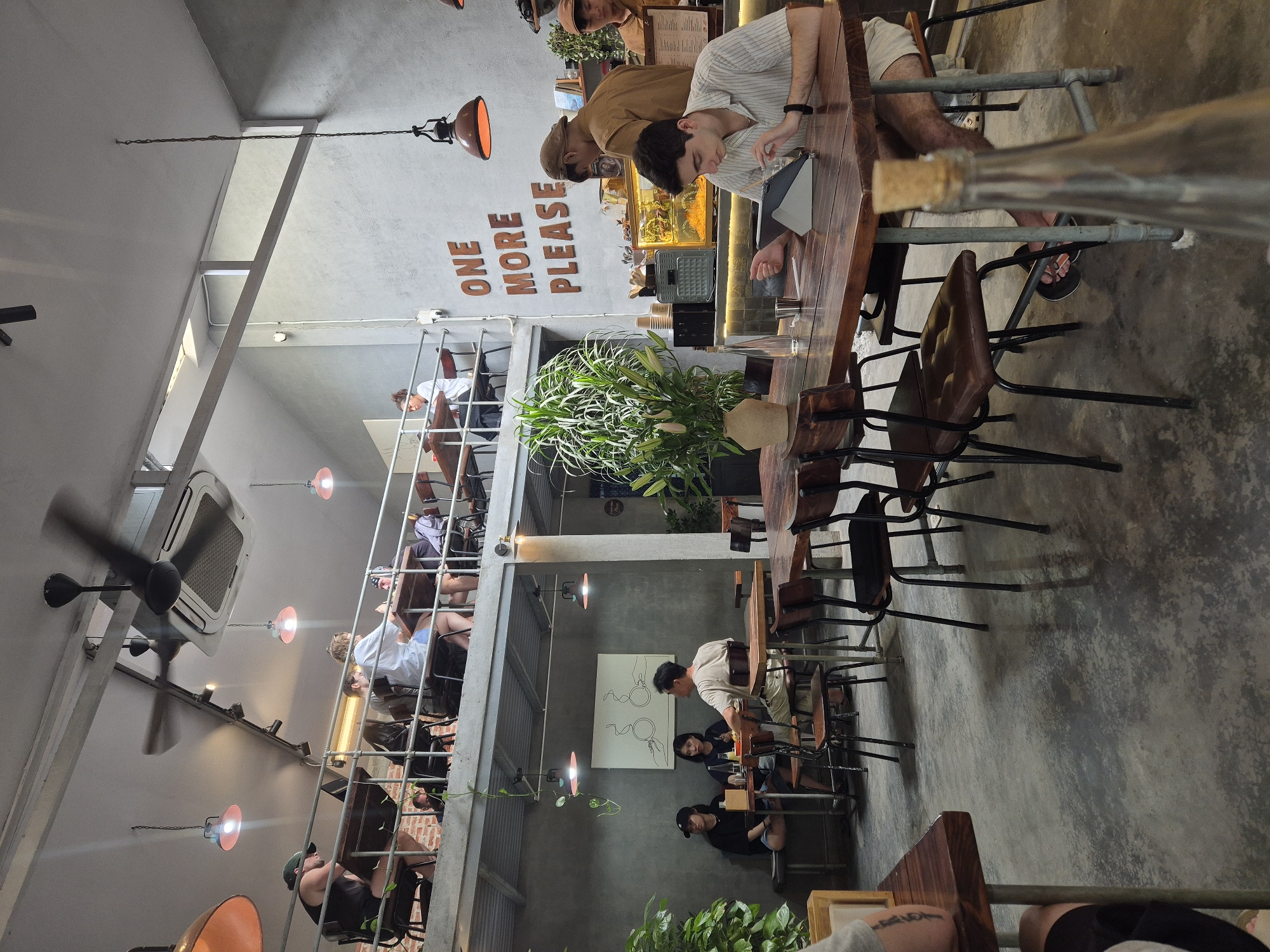 The image depicts the interior of a multi-level cafe in Đà Nẵng, Vietnam. On the upper level, several patrons are seated at tables, some appearing to work on laptops. The lower level features a long communal table where a young man is focused on a tablet, a bottle of water, and a small metal cup. A large potted plant with white flowers is situated near the center of the lower level. Behind the communal table, a counter area is visible with staff members and a display case. A prominent wall decoration reads "ONE MORE PLEASE" in large wooden letters. The cafe's ambiance suggests a modern, industrial aesthetic with exposed concrete walls, metal structures, and warm lighting from pendant lamps. The floor is a polished concrete.