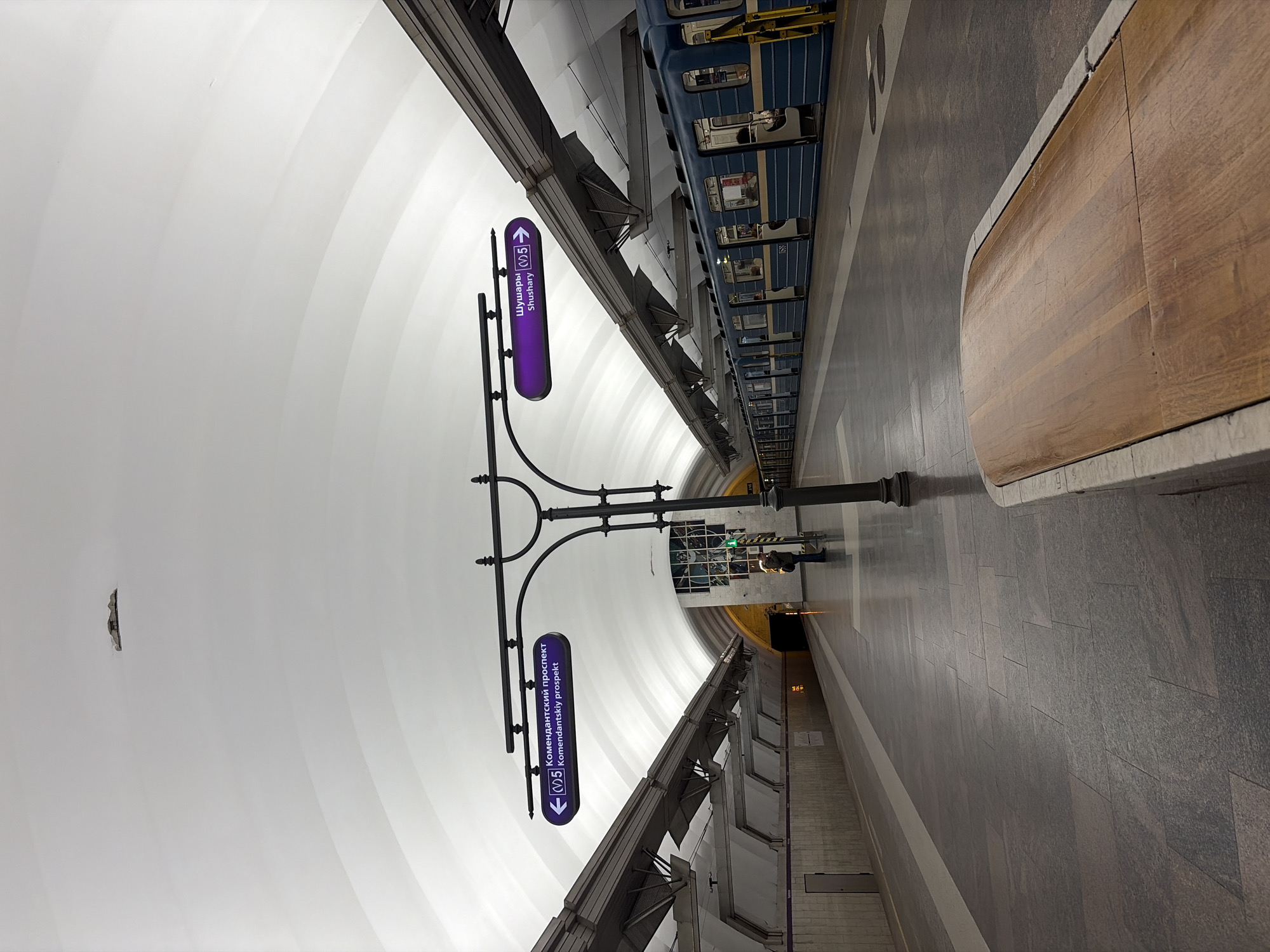 The image shows the Shushary metro station in Saint Petersburg, Russia. The view is along the platform with a train visible to the right. There are two purple signs with directions in Russian. A person stands on the platform in the distance. The ceiling is white and arched. The floor is made of grey tiles. A wooden bench is next to the platform edge.