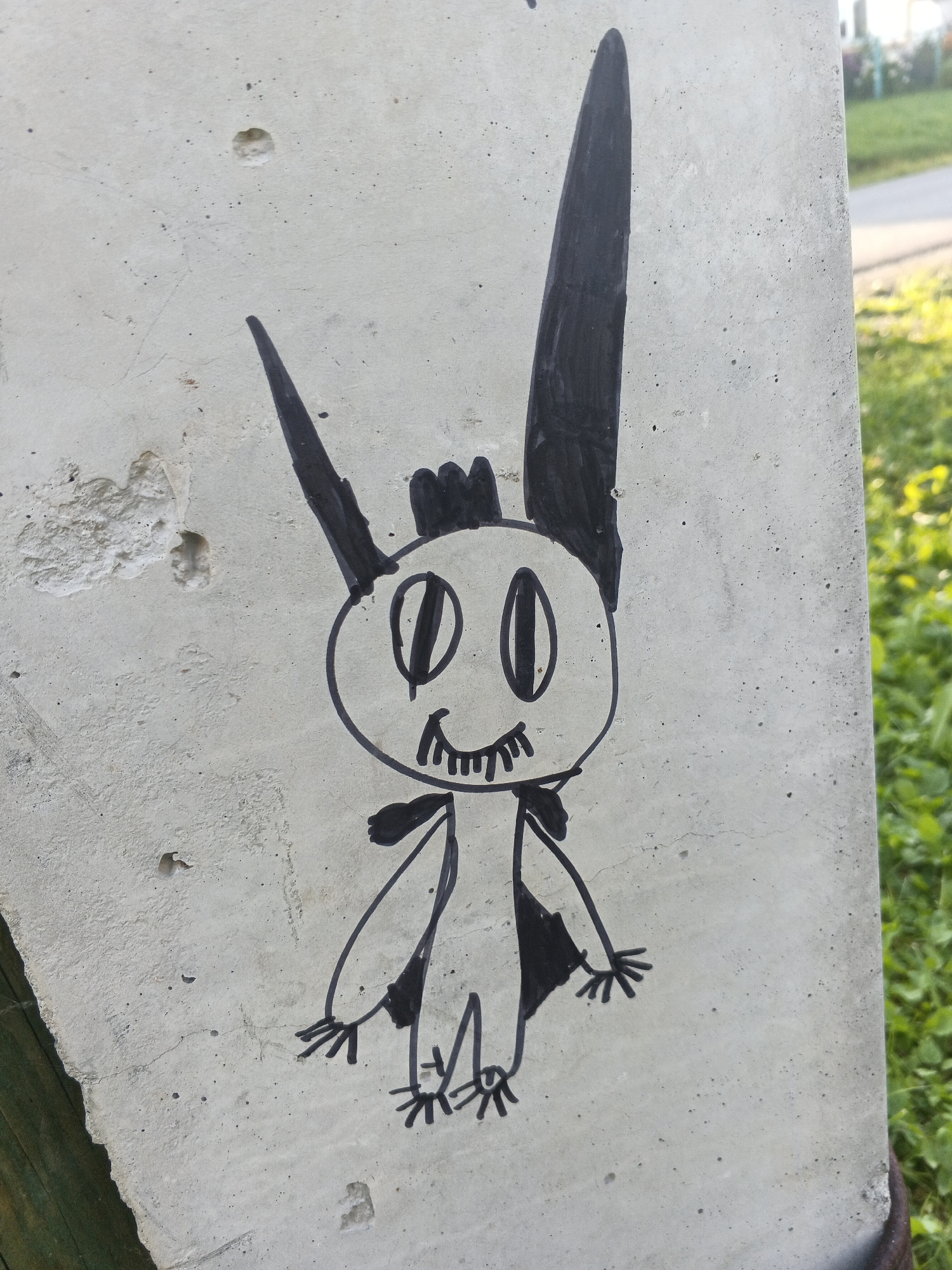 A black marker drawing of a cartoon monster is on a concrete surface. The monster has a round head with two large oval eyes, a wide mouth with jagged teeth, and what appear to be two pointy ears and a crown-like structure on its head. It has stick-like arms and legs with stick fingers and toes, and two triangular cape-like shapes attached to its shoulders. The concrete surface is rough and has some cracks and small holes. In the background, out of focus, is green grass and a hint of a path or road. The image is a close-up, focused on the drawing. There is no visible indication of the specific location in Medvedyovka, Russia.