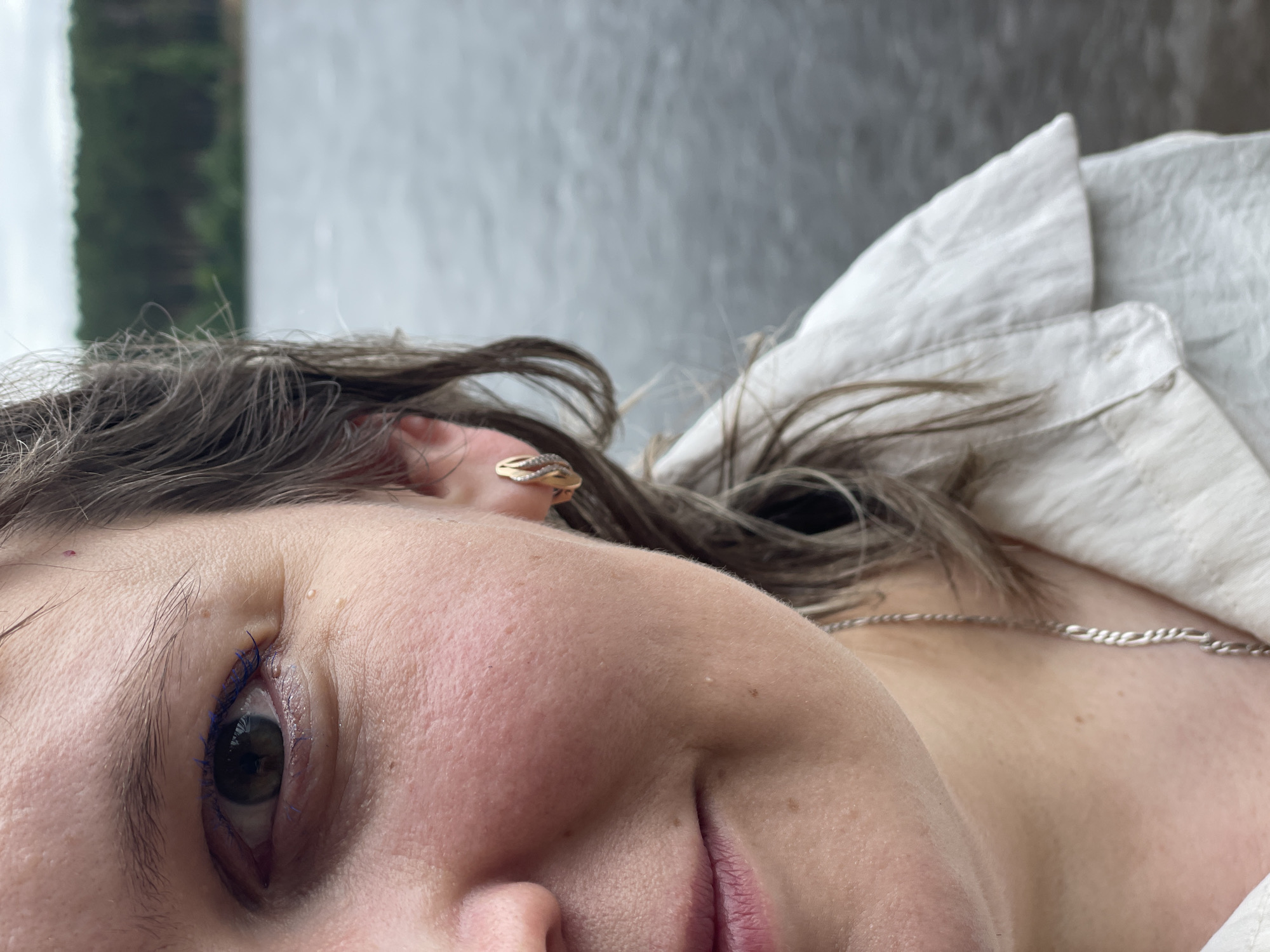 A close-up, profile view shows a woman looking to her left, her face is partially obscured by her dark brown hair. She is wearing a light-colored shirt and a silver chain. A gold earring with diamonds adorns her left ear. The background is a blurred body of water, likely the Kama River, with a treeline visible in the distance. The image was taken outdoors.