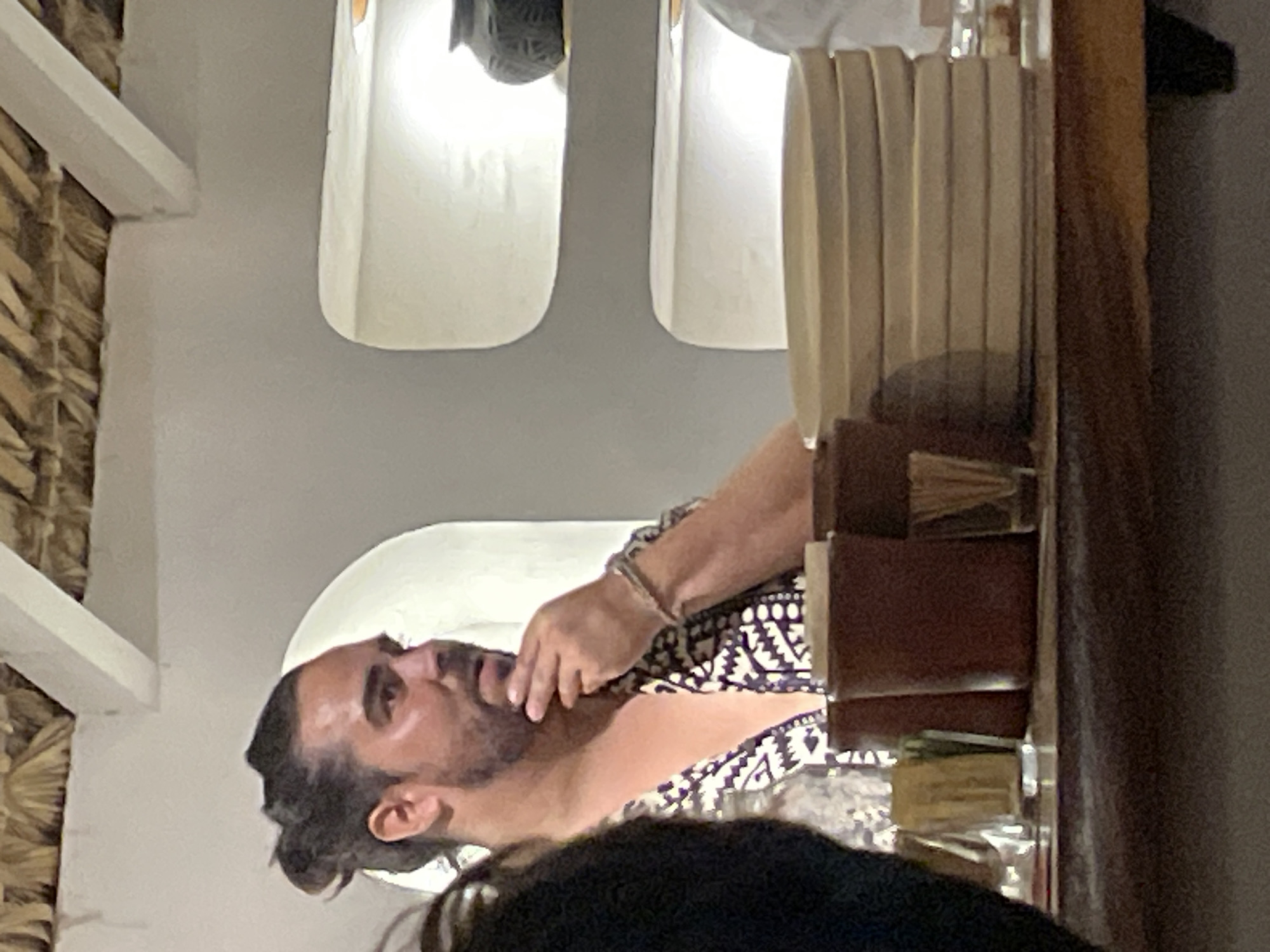 A man with a beard and hair in a bun sits at a table in an indoor setting in Brisas de Zicatela, Mexico. He is wearing a patterned shirt and a bracelet. He is looking at something or someone at the table. The table is set with items that are partially obscured, including what appears to be plates or placemats. The architectural elements around him consist of whitewashed walls, arched windows, and exposed beams.