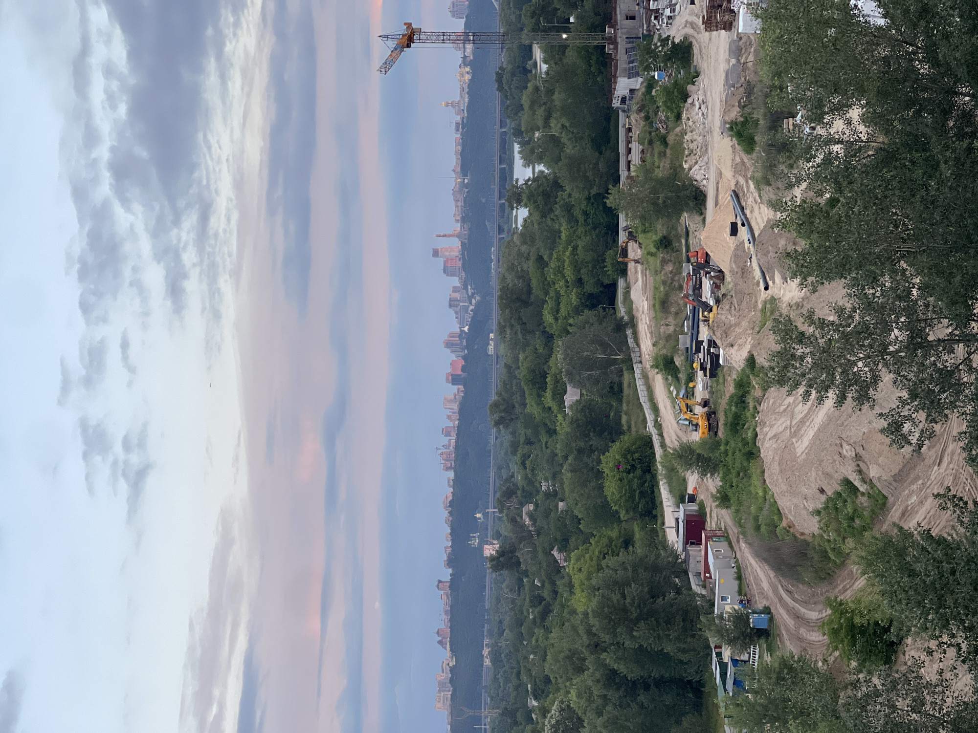 This is an aerial, vertical shot of a construction site in Kyiv, Ukraine, with a distant cityscape visible in the background. The sky is a soft blend of blue, pink, and gray, suggesting dawn or dusk. In the foreground, a dirt road winds through a construction area with piles of sand and gravel. Various construction vehicles, including excavators and a crane, are present. Several modular buildings, possibly site offices, are situated near the road. Further back, dense green trees dominate the landscape, interspersed with some houses. Beyond the trees, a hazy cityscape with numerous buildings and a bridge crossing a body of water stretches across the horizon. The overall scene depicts urban development against a natural and sky backdrop.