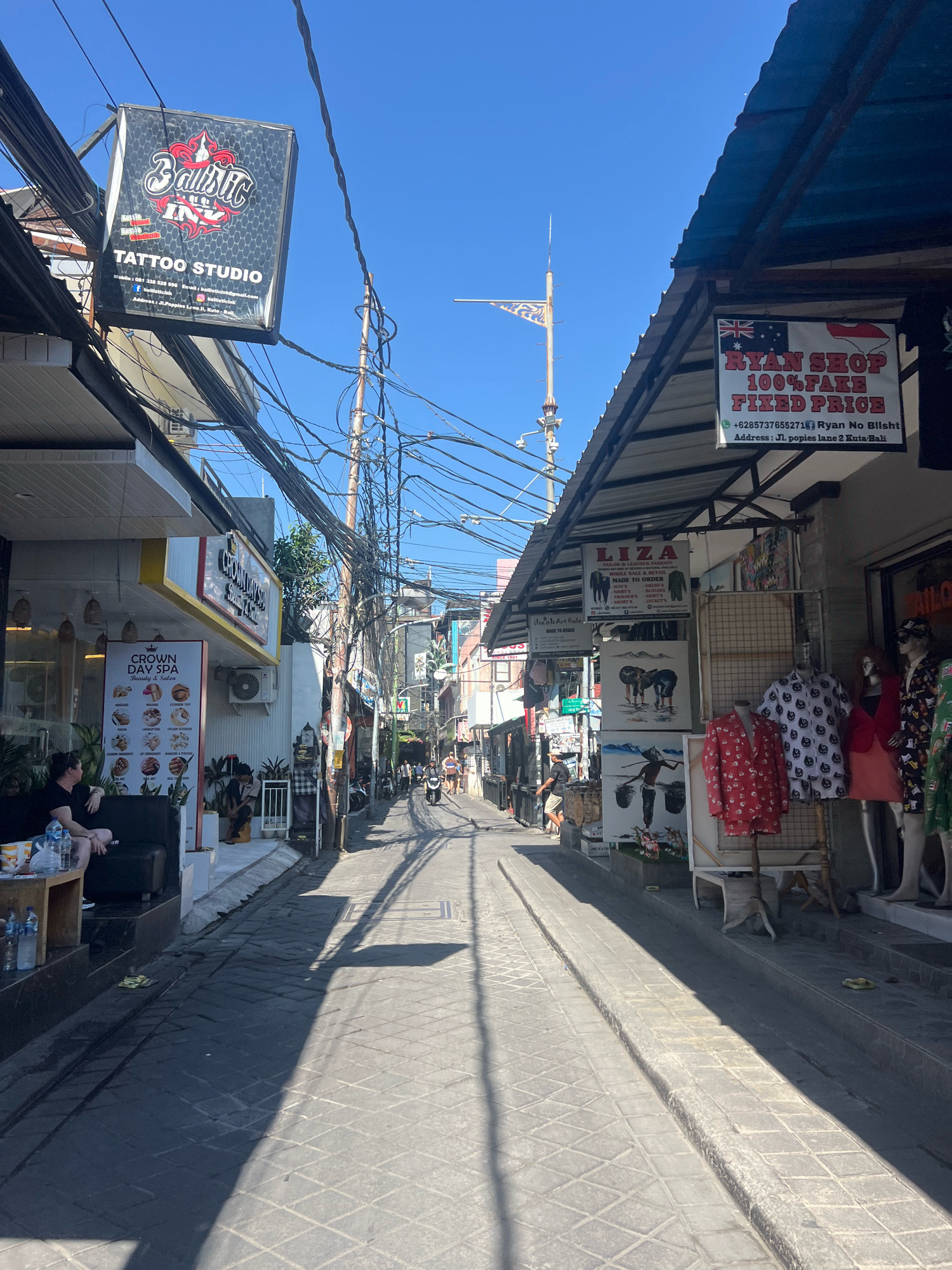 The image depicts a street scene in Kuta, Indonesia, likely a tourist area given the presence of shops and signs. A narrow, paved street runs from the foreground into the distance, flanked by businesses on both sides. The architecture appears to be a mix of single and multi-story buildings, many with open-fronted shops displaying goods. Overhead, a dense network of electrical wires crisscrosses the sky.
On the left side of the street, a shop called "Ballistic Ink Tattoo Studio" is prominently visible with its signage. Further down, "Crown Day Spa" is advertised with a list of services. Several individuals are present: one person sits on a black couch outside the spa, and a few others are seen walking or standing further down the street.
The right side of the street features several shops. One sign reads "RYAN SHOP 100% FAKE FIXED PRICE." Below this, another sign advertises "LIZA" for "SUITS, SHIRTS, TROUSERS, etc., MADE TO ORDER." Several mannequins are displayed in front of a shop, dressed in patterned clothing. Artwork depicting elephants and a figure with baskets is also displayed outside a shop.
The street itself is paved with grey cobblestones, and the harsh sunlight casts strong shadows, indicating it is daytime. The overall impression is a bustling commercial street catering to tourists.