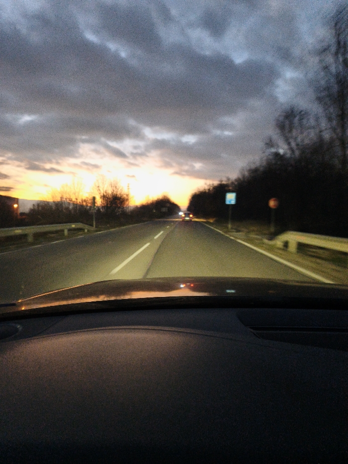 The image depicts a roadside scene at dusk or sunset in Grigorevo, Bulgaria, viewed from inside a vehicle looking forward through the windshield. The lower quarter of the frame shows the dark dashboard of the vehicle, with a reflective strip at the base of the windshield. The two-lane asphalt road ahead is illuminated by the vehicle's headlights, casting a yellowish light. A solid white line marks the left edge of the road, and a dashed white line separates the lanes. A metal guardrail parallels the road on the immediate left, with another partially visible on the right. The horizon displays a vibrant sunset with strong orange and yellow hues, while the sky above is dominated by dark, heavy grey and purplish clouds. Bare trees and shrubs line both sides of the road. In the distance, several vehicles are visible, characterized by their illuminated headlights in the opposing lane and taillights in the same direction. Two road signs are discernible on the right side of the road: a rectangular blue sign with white iconography and a circular red and white sign further ahead.