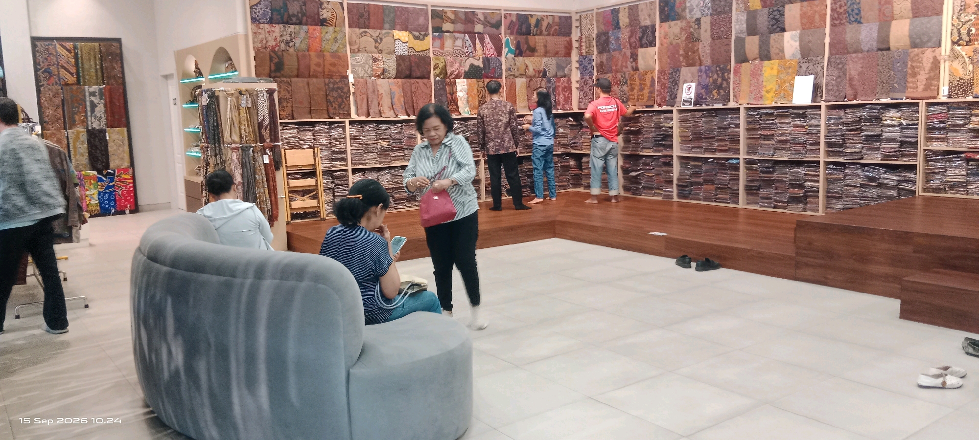 The image depicts the interior of a batik shop in Surakarta, Indonesia. In the foreground, a light grey upholstered sofa is visible, with two individuals seated on it. One person, viewed from behind, is wearing a light blue hooded jacket and appears to be looking at their phone. To the right of the sofa, a woman wearing a striped shirt and black pants stands holding a red purse. She is looking downwards. Behind her, a man in a red t-shirt and jeans stands with his hands on his hips. Further in the background, shelves are filled with folded batik fabric. Another man in a patterned long-sleeved shirt and dark pants stands near the shelves, facing away from the camera. The walls are covered with various batik patterns displayed as hanging textiles and on shelves. The floor is tiled in the foreground and transitions to wooden platforms with tiered steps in the background. The shop appears to be well-stocked with batik merchandise.