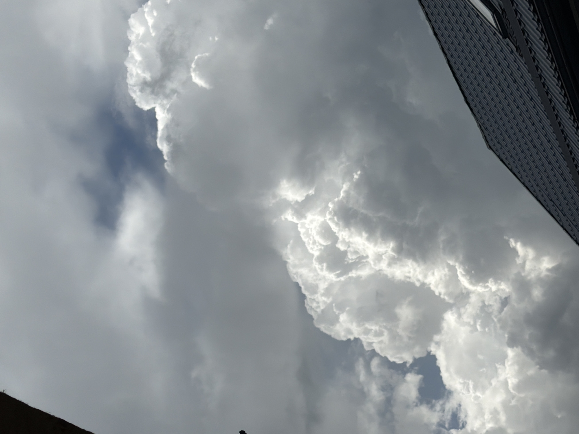 The image shows a view of the sky in Rossiyskiy, Russia. Large, billowing clouds dominate the frame, with varying shades of grey and white. Some patches of blue sky are visible between the clouds. The corner of a building with a textured exterior is visible in the upper right of the image. The angle suggests the photo was taken looking upwards.