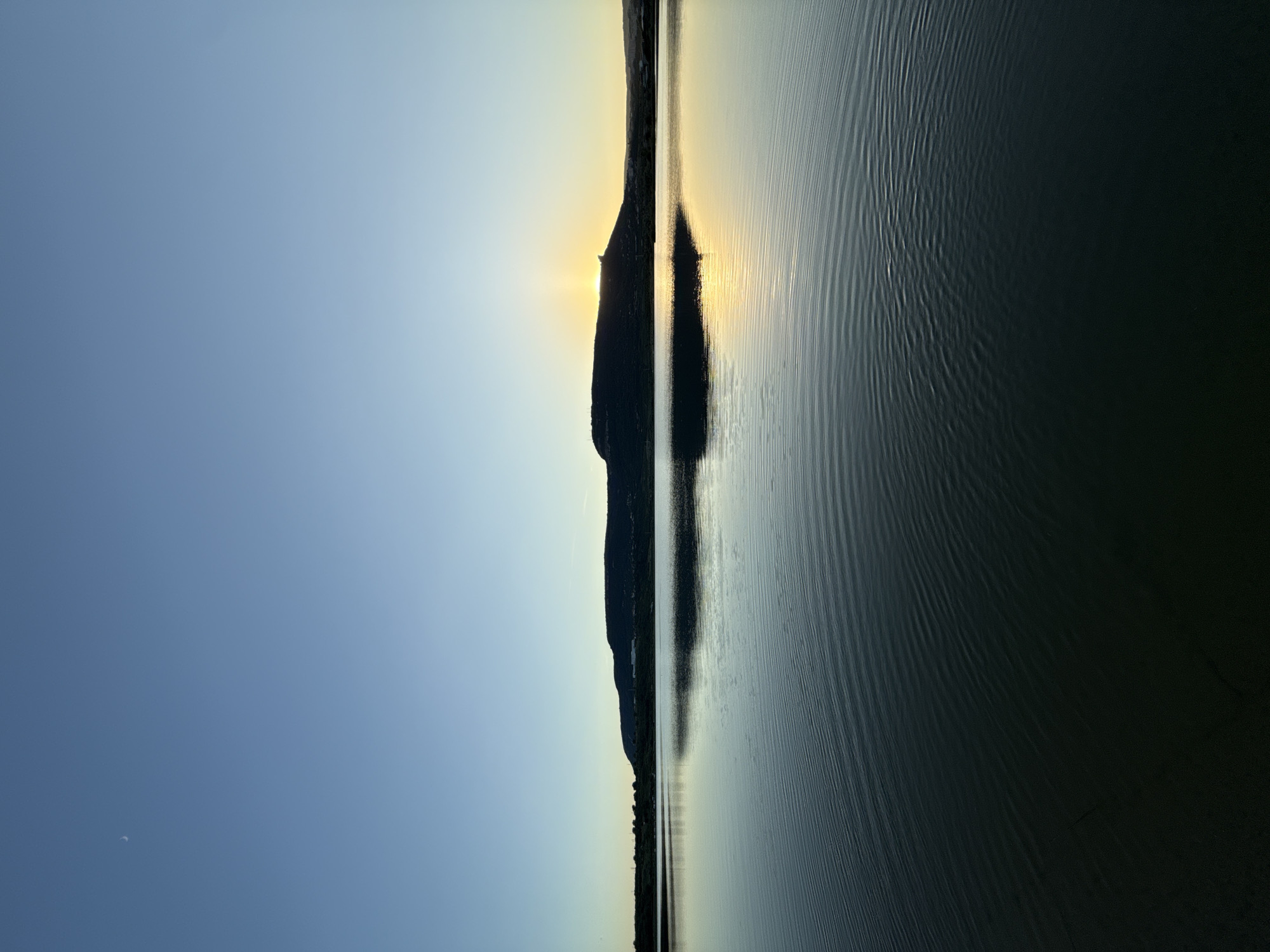 The image depicts a tranquil lake at dawn or dusk, with a crescent moon visible in the clear blue sky. The water is calm, reflecting the soft hues of the sky and the silhouette of a distant hill. The sun is just beginning to peek over the horizon, casting a warm golden glow on the landscape. The scene evokes a sense of peace and serenity. The specific location in Shumen, Bulgaria, cannot be determined from the image alone, as it lacks any identifying landmarks or context. The image appears to be a landscape photograph, taken without any human presence or activity.