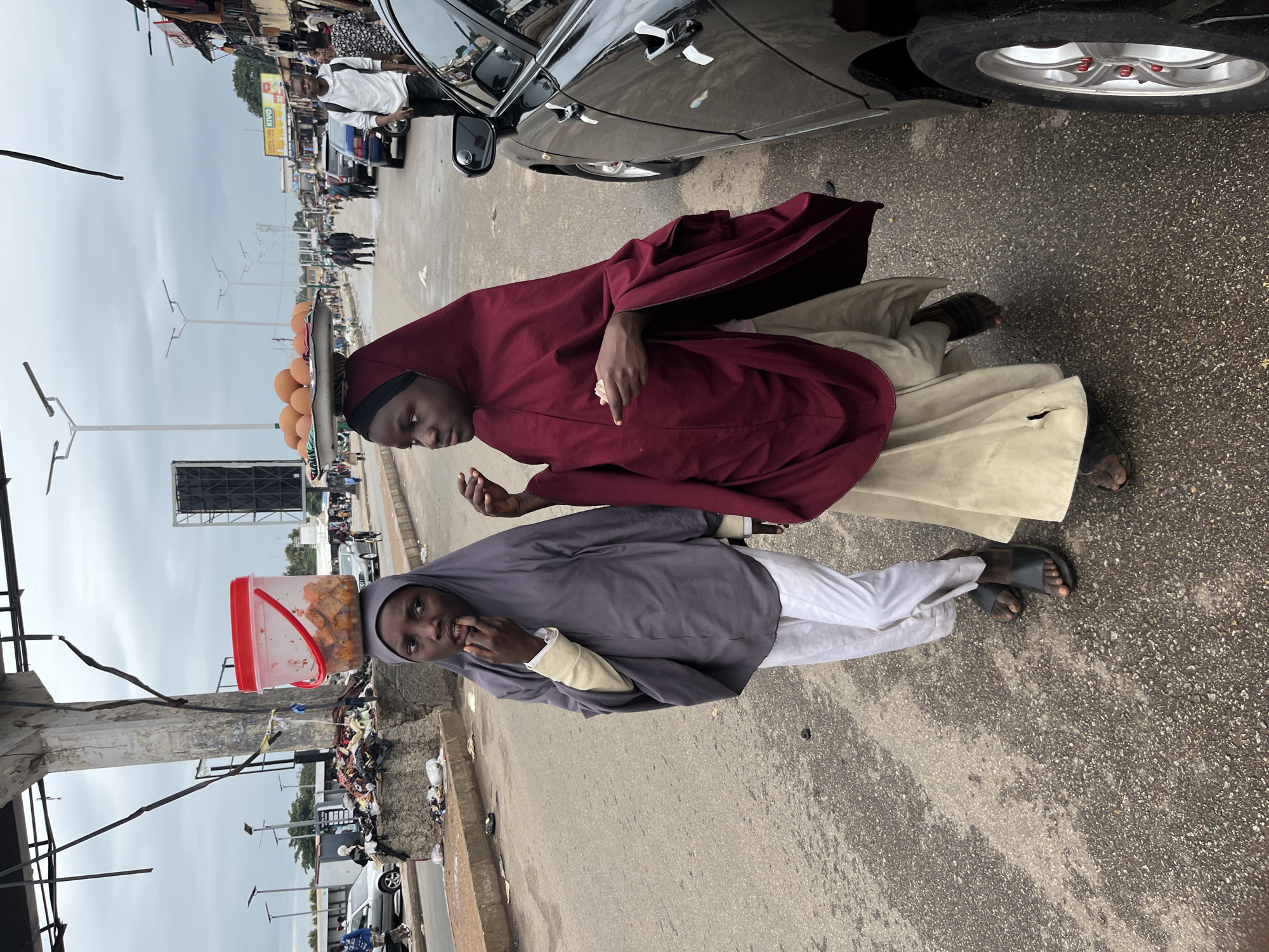 In Jos, Nigeria, two young women walk along a paved road, one carrying a red plastic bucket filled with fried food on her head, and the other balancing a tray of eggs. Both are dressed in Islamic attire, with one wearing a grey hijab and white pants, and the other a burgundy hijab and a beige skirt. They are walking alongside a black car, with other pedestrians and vehicles visible in the background. The environment appears to be a bustling urban area, with street vendors and a cloudy sky.