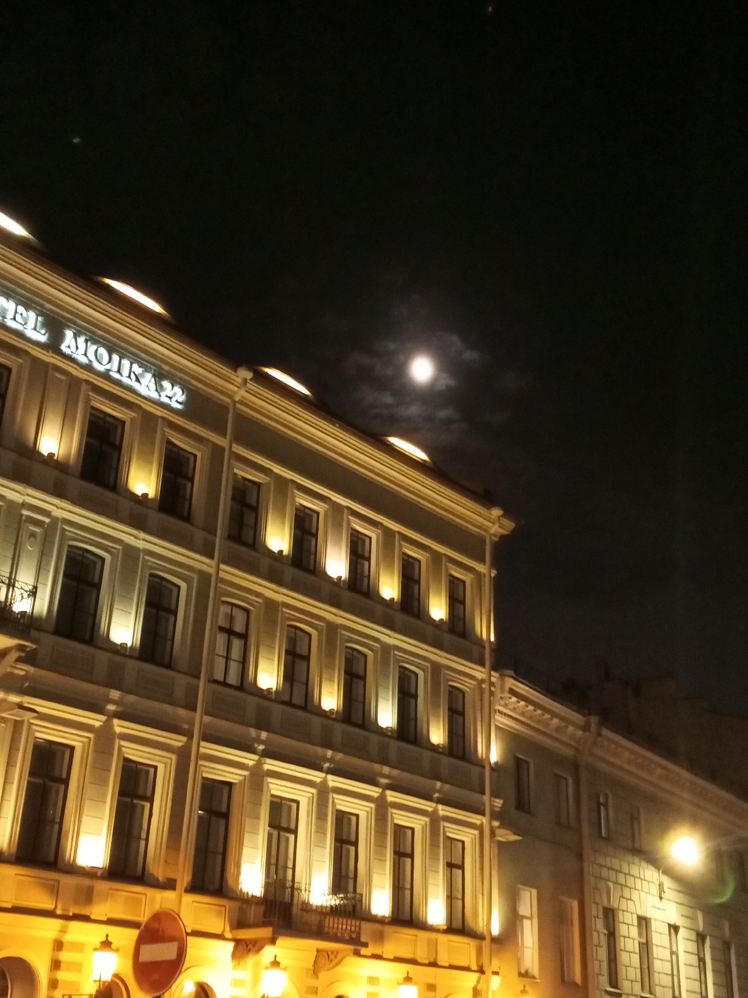 At night, the image shows the Hotel Moika 22 in Saint Petersburg, Russia. The illuminated facade of the hotel contrasts against the dark sky, where a full moon shines. The hotel's signage, "HOTEL MOIKA 22," is lit up atop the building. Streetlights illuminate the lower part of the building and the street, creating a warm glow. A "no entry" road sign is visible in front of the hotel. Other buildings are also visible in the distance.