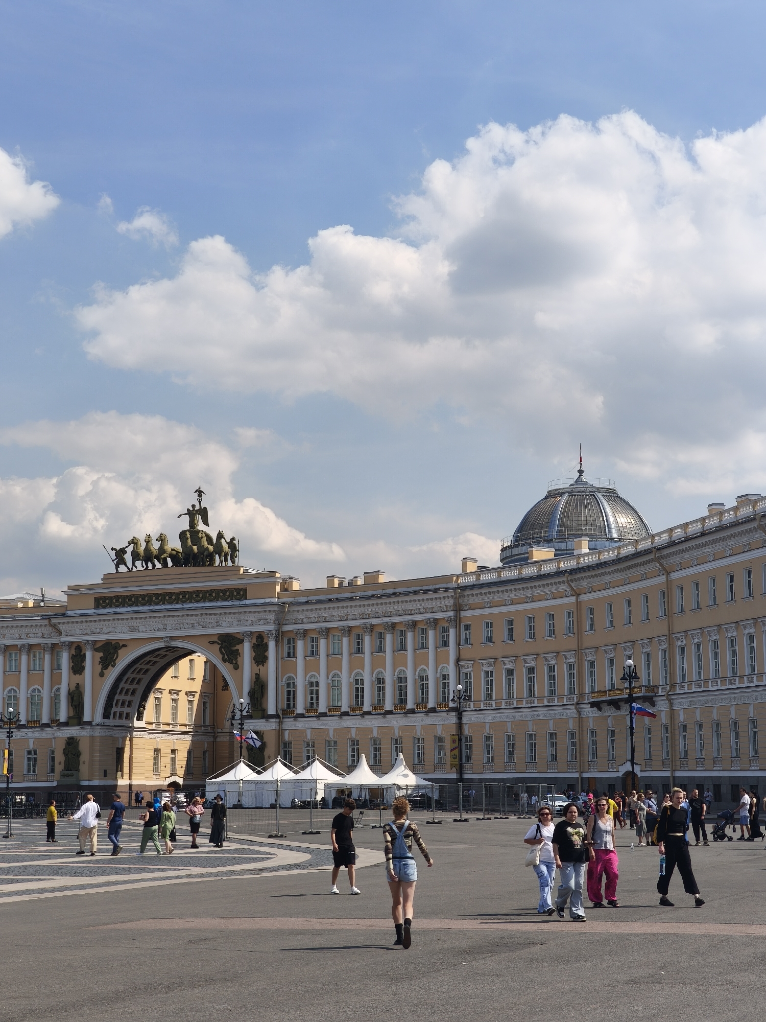The image depicts the Palace Square in Saint Petersburg, Russia, on a sunny day with scattered clouds. A grand neoclassical building with an arched entrance dominates the background, featuring a chariot sculpture on its apex. To the right, a curved section of the building is visible, crowned by a glass dome. In the foreground, a paved square is populated by numerous individuals walking and standing. Several white tents are set up in the middle ground, surrounded by temporary fencing. The scene captures a moment of everyday activity in a significant historical and architectural landmark of the city.