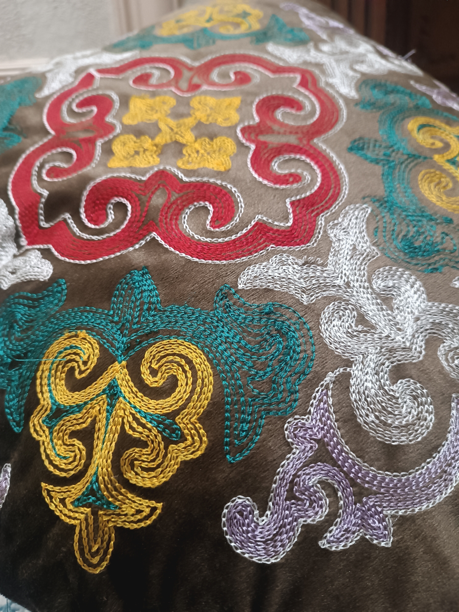 The image is a close-up shot of a brown fabric, possibly a cushion or tapestry, adorned with intricate embroidery. The embroidery features a repeating pattern of swirling, organic shapes in various vibrant colors including red, yellow, teal, white, and lavender. The background fabric has a soft, possibly velvet, texture. The lighting suggests an indoor setting, with the focus on the detailed craftsmanship of the embroidery. There are no people visible in the image. The image is a still photograph of a textile object. It is not possible to determine the specific location of Myrza-Ake, Kyrgyzstan from this image.