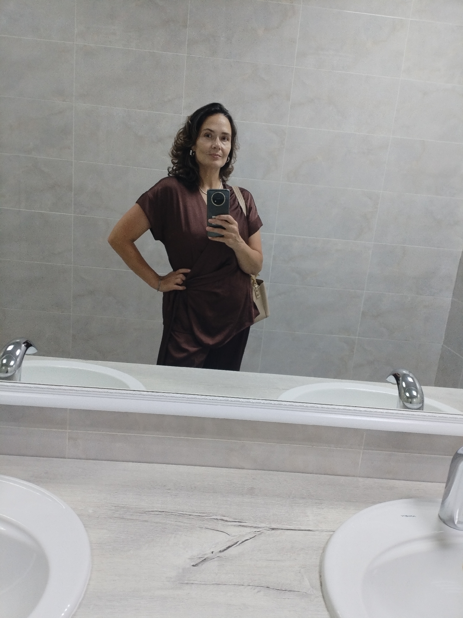 A woman stands in front of a bathroom mirror, taking a selfie with her phone. She has dark, wavy hair and is wearing a brown, draped top and dark pants. A light-colored purse is slung over her shoulder. The background is a tiled wall and the reflection shows a bathtub and two sinks with chrome faucets. The overall scene is a clean, modern bathroom. The photo was taken in Vinogradny, Russia.