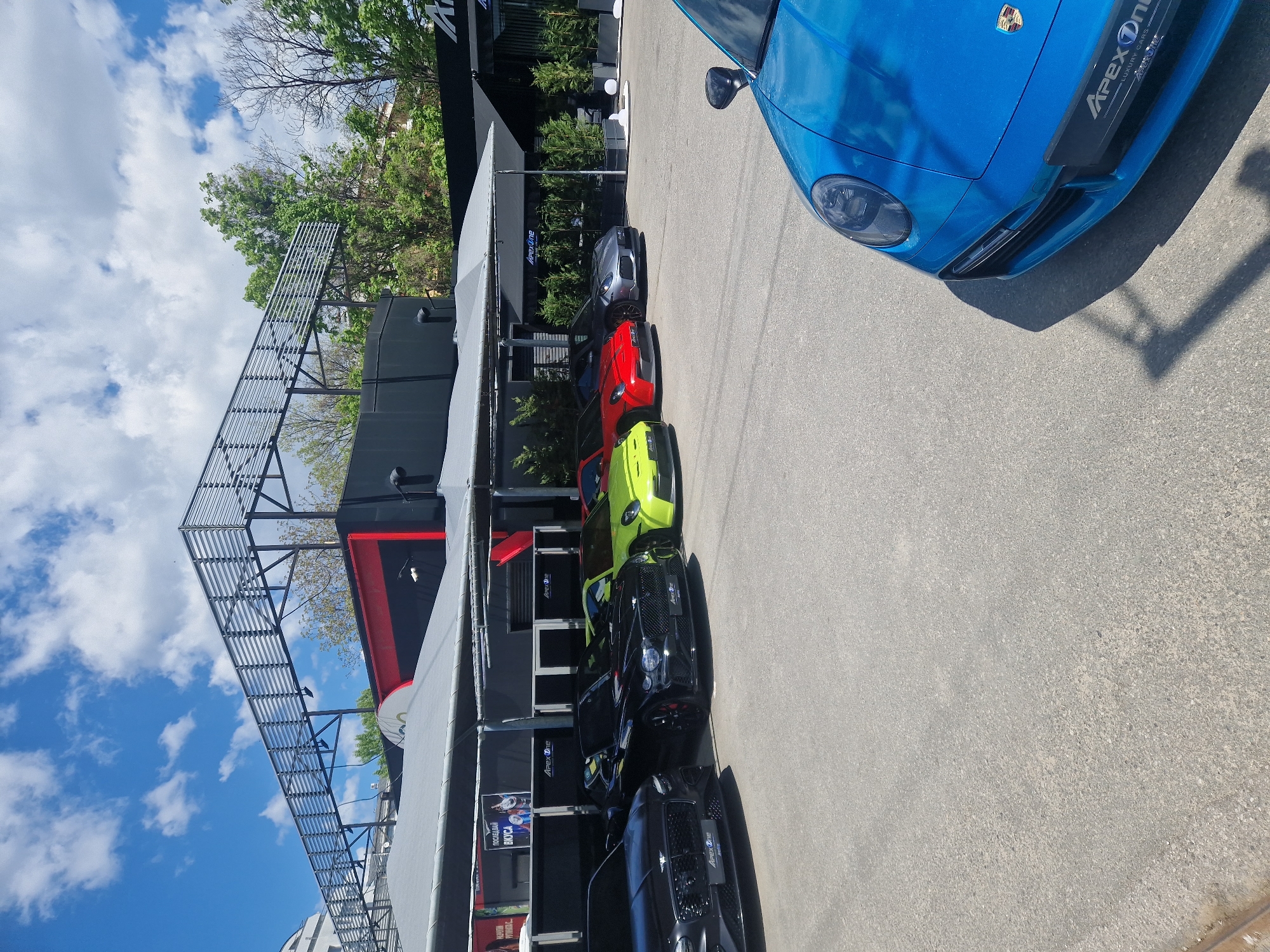 The image shows a line of luxury cars parked next to a building with a metal structure on top. The cars are in various colors, including blue, gray, red, and green. The building has an "Apex One" sign on it. The sky is blue with some clouds. The location is Unknown, Unknown.