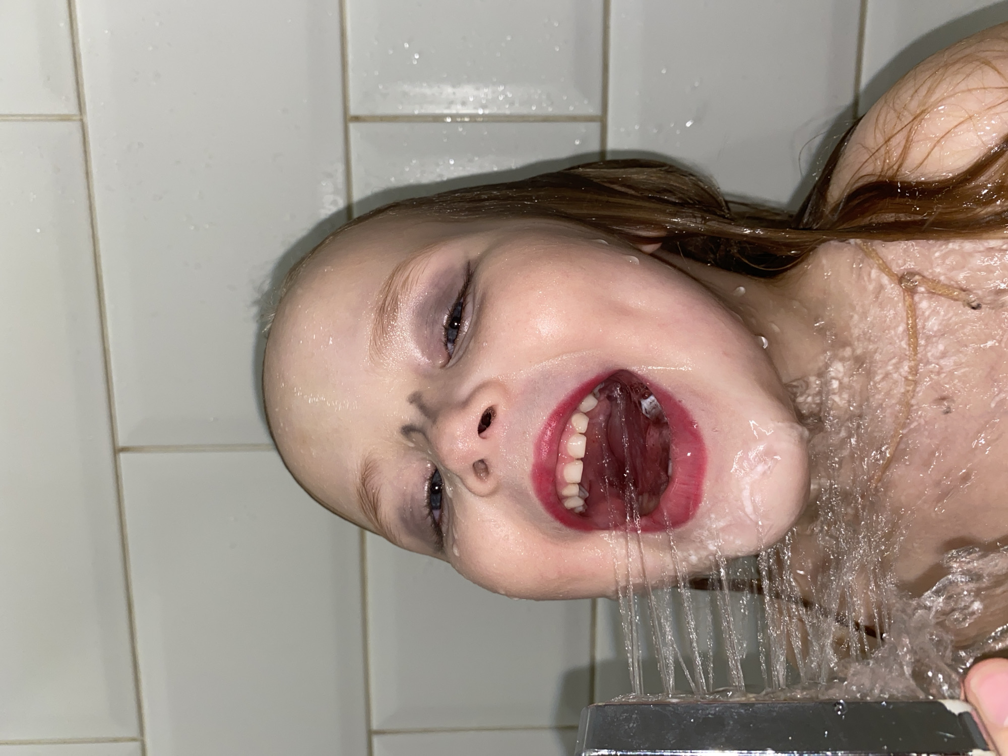 A close-up, rotated image captures a young child's face and upper body in a shower in what appears to be Votkinsk, Russia. The child's head is tilted to the left, with her long, wet brown hair clinging to her right shoulder and upper back. Water streams forcefully from a chrome shower head, hitting her face and pouring directly into her widely opened mouth. Her lips are adorned with red lipstick, which shows signs of being washed away by the water, revealing her tongue and white teeth.
Dark smudges are visible on her nose and under her left eye, possibly makeup or dirt, adding to a playful or slightly disheveled appearance. Her blue eyes are open and gaze towards the viewer, conveying an expression of playful surprise or exhilaration from the water. Her skin is visibly wet, with numerous water droplets glistening on her face and shoulders.
The background consists of plain white rectangular tiles with dark grout lines, typical of a bathroom shower stall. Water droplets are also scattered across the tiled wall. The image captures an energetic moment of the child interacting directly with the shower water. No text is visible in the media file.