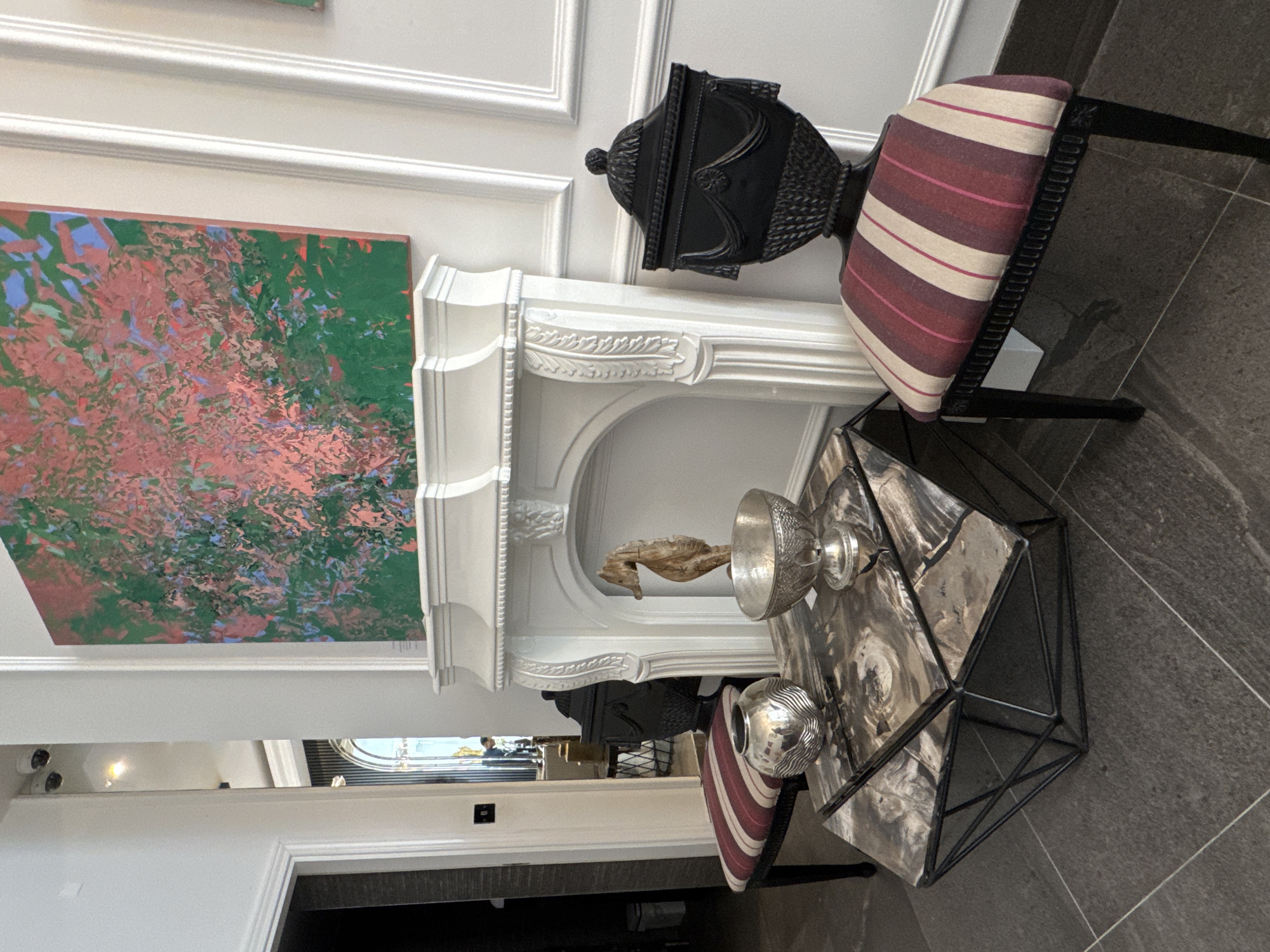 This is a medium shot, eye-level, interior photograph of a decorative fireplace surround and its adjacent furnishings, taken inside a modern, well-lit room in London, United Kingdom. The fireplace is white, ornate, and features a carved mantelpiece and decorative trim. Above the fireplace, a large, abstract painting with bold strokes of green, pink, and blue hangs on a white paneled wall. In front of the fireplace, a black metal geometric coffee table with a marble-like top displays two silver decorative bowls and a carved wooden seahorse sculpture. To the left of the coffee table, a chair with a black frame and a striped seat cushion in shades of red, pink, and beige sits on a dark grey tiled floor. To the right of the coffee table, a matching chair with a black frame and a similar striped seat cushion is visible. The overall impression is one of sophisticated interior design.