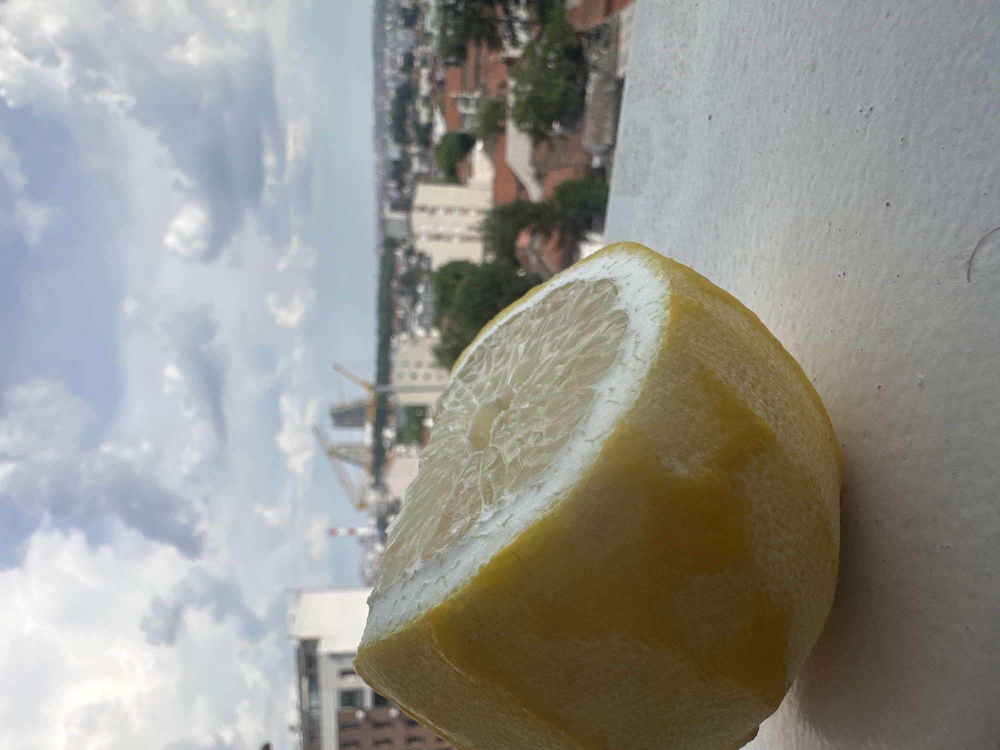 This media file captures a close-up of a cut lemon on a textured surface. The lemon's bright yellow rind and juicy, segmented interior are clearly visible, with the rind showing a slightly rough texture. In the background, out of focus, is a cityscape viewed from a high vantage point. Buildings with red roofs and some taller, modern structures are discernible, interspersed with greenery. Construction cranes are also visible in the distance, suggesting ongoing development. The sky above is filled with dramatic, fluffy white clouds against a pale blue backdrop, indicating a daytime scene, likely late morning or afternoon, with fair weather. The scene's orientation is such that the lemon is in the foreground, angled upwards, while the cityscape stretches out beyond it. There is no visible text in the media file. The setting appears to be an elevated outdoor location, possibly a balcony or rooftop, in Belgrade, Serbia, given the architectural style and the presence of construction cranes.
