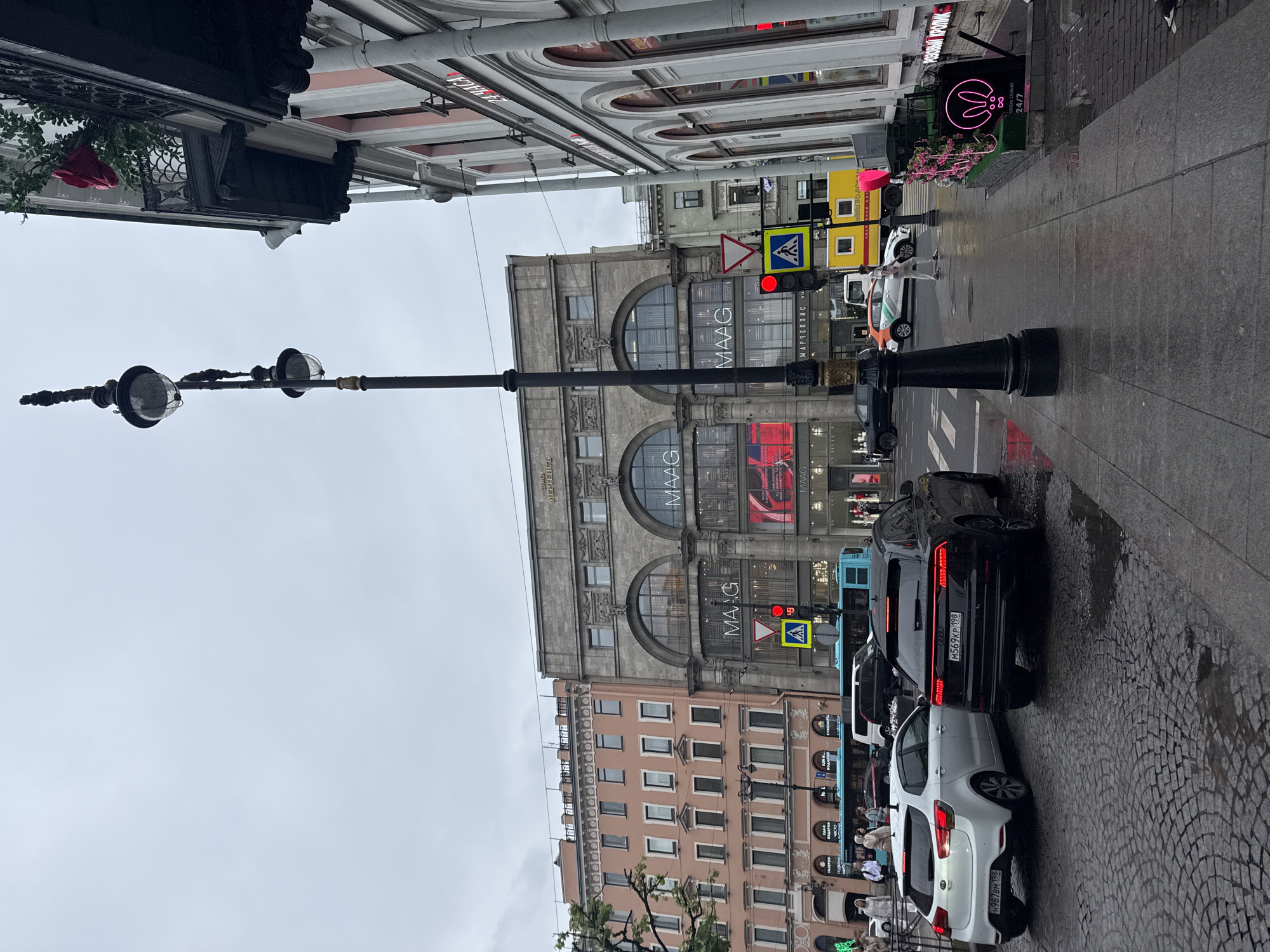 This image depicts a street scene in a city. Several cars are parked along a cobblestone street, which appears wet, suggesting recent rain or a damp environment. In the background, large, multi-story buildings line the street, displaying various signage. One prominent building features the "MAAG" logo multiple times, along with Russian text. Another building has a glowing pink bunny logo and the text "РОЗОВЫЙ КРОЛИК" and "24/7". Traffic lights and pedestrian crossing signs are visible, indicating an active intersection. A few people are seen walking on the sidewalk. The sky is overcast, suggesting a cloudy or possibly late afternoon/early morning time. The overall mood is ordinary urban life.