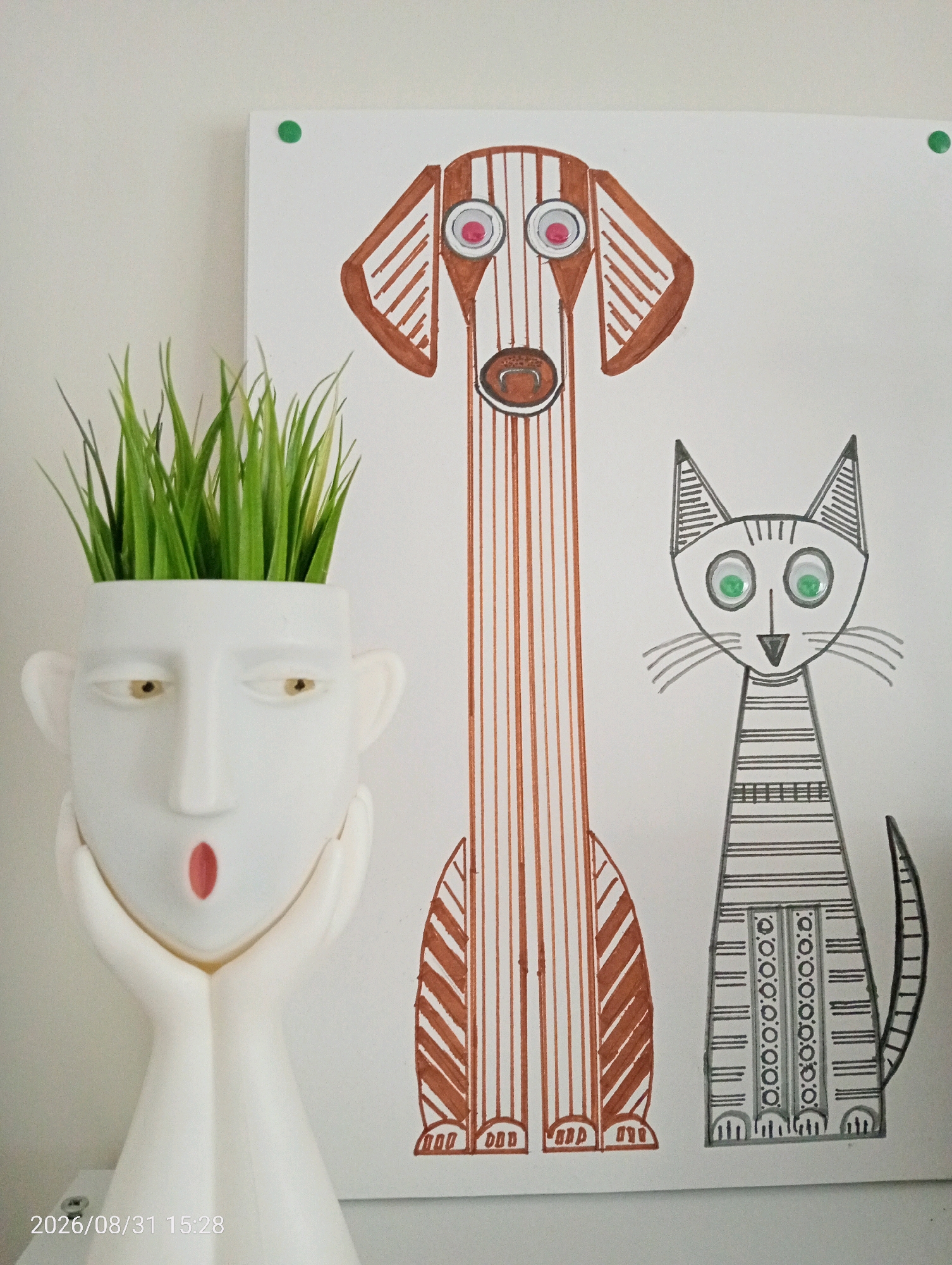 A white ceramic planter in the shape of a stylized human head holding its face with hands contains artificial green grass. To the right of the planter, a drawing on white paper depicts a long, brown and white striped dog with wide eyes and a red tongue, and next to it, a gray and white striped cat with green eyes and whiskers. The drawing is affixed to a white wall. A timestamp in the bottom left corner reads "2026/08/31 15:28". This image was taken in Silifke, Turkey.