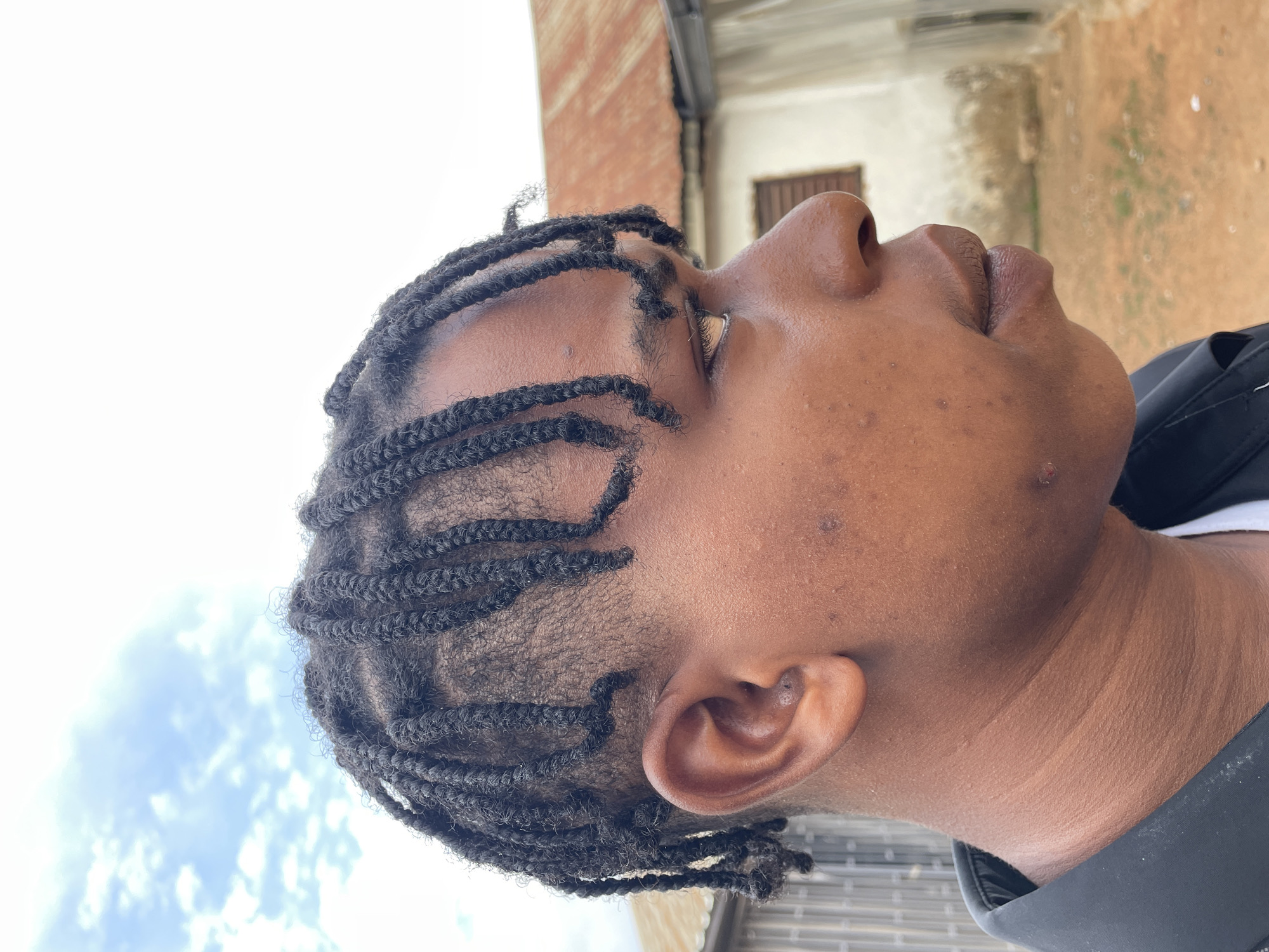 The image captures the right profile of a person with dark skin, their head tilted upwards and slightly back, looking towards the sky. They have tightly woven black braids (cornrows or similar style) covering their scalp. The person's right eye is partially visible, appearing to gaze upwards, with a calm or neutral expression. Their nose is prominent, and their mouth is closed. Natural creases and folds are visible on their neck, which is supported by a dark-colored garment, possibly a jacket collar, over a hint of a white undershirt.
The background is divided, with a bright, largely clear sky featuring scattered white clouds on one side, and an outdoor, somewhat rustic environment on the other. This environment includes what appears to be a building with a reddish-brown corrugated metal roof and lighter-colored walls, possibly indicative of a residential or commercial structure. The ground beneath suggests an earthen or dusty area. The bright, natural lighting indicates it is daytime. The scene takes place in Jos, Nigeria. No visible text is present in the image.