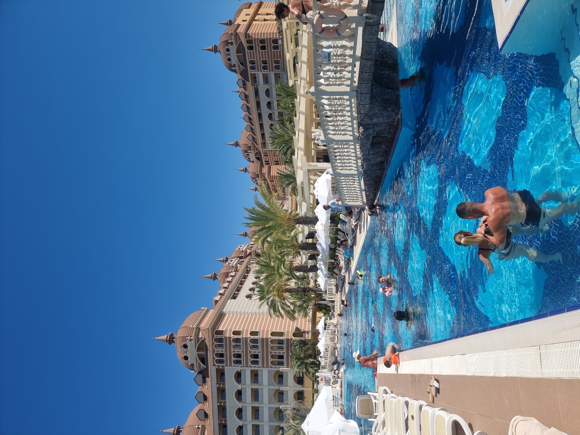 The image shows a large outdoor swimming pool with crystal clear blue water, decorated with a mosaic pattern on the bottom. Several people are enjoying the water, some swimming, some wading, and some relaxing. In the background, a grand, multi-story hotel with traditional architecture and domed roofs is visible, surrounded by palm trees. The sky is a clear, bright blue, indicating a sunny day. A white pedestrian bridge arches over a section of the pool, connecting two parts of the hotel or its grounds. Sun loungers and white umbrellas are arranged around the pool deck, suggesting a resort setting. The overall scene is one of leisure and vacation in a warm climate. This scene appears to be at a resort located in Manavgat, Turkey, known for its extensive hotel complexes and proximity to natural attractions.