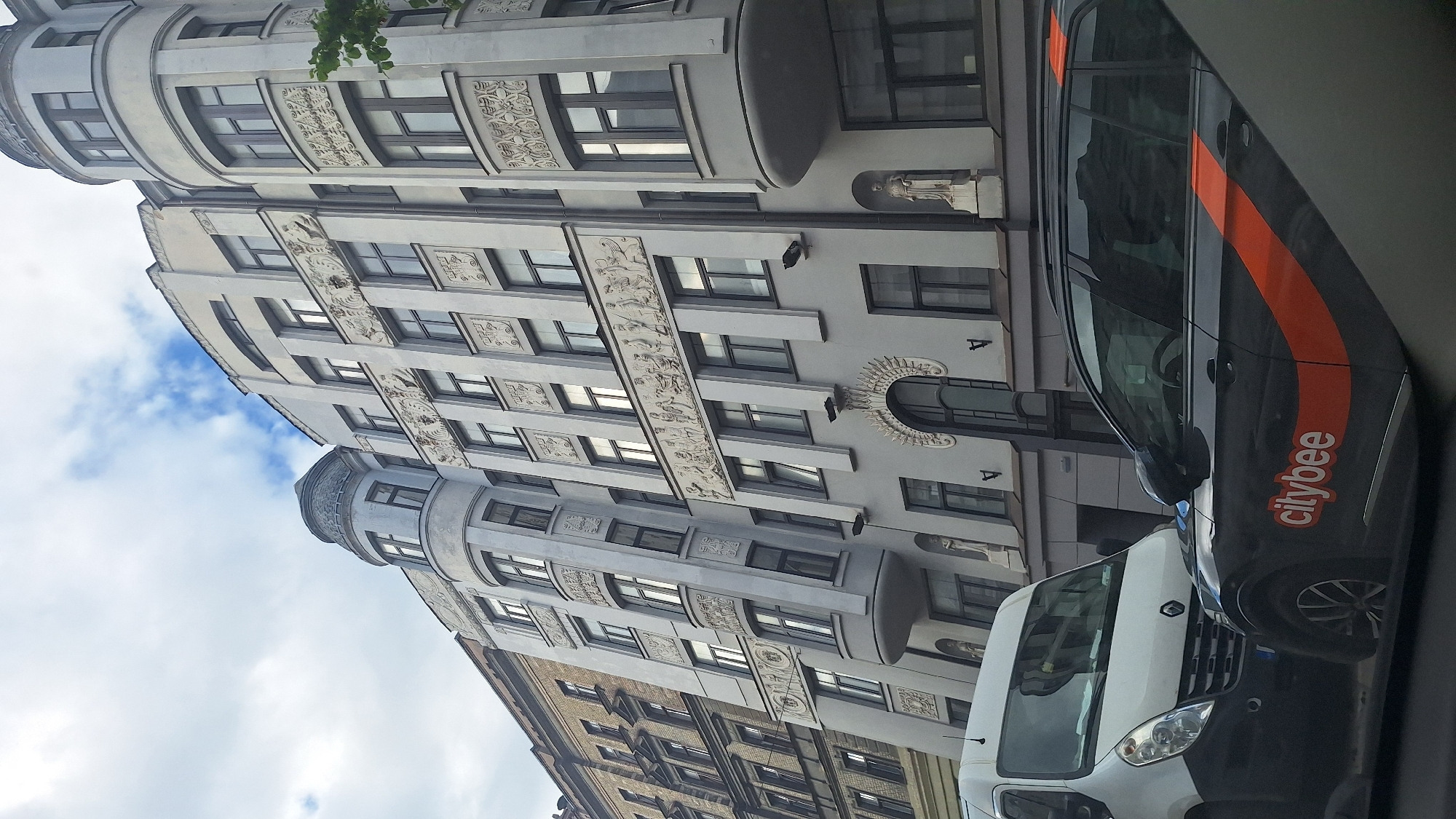 This is an eye-level shot of an Art Nouveau building in Riga, Latvia. The building is ornate with decorative reliefs of figures and geometric patterns along the facade. The main part of the building is light grey with dark framed windows. There are rounded tower-like sections on the left and right sides of the building. To the left of the Art Nouveau building is a narrower, older building with a yellow facade. In the foreground, a black car with an orange stripe and the logo "citybee" is visible. A white van is parked next to it. The sky is overcast with some blue sky peeking through. The image is likely taken from another vehicle, as indicated by the reflections in the car window and the cropped perspective.