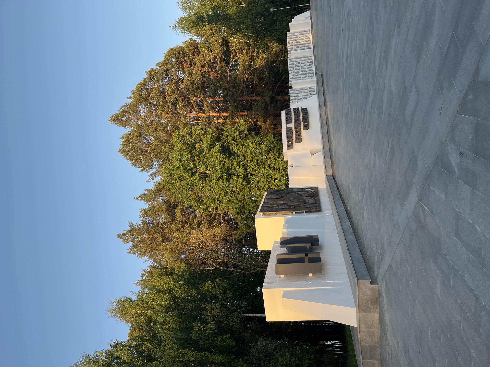 The image depicts a monument in Chaikovsky, Russia. The monument consists of white, tiered rectangular blocks with dark inscriptions and relief sculptures. A paved area extends from the monument, constructed of grey rectangular slabs. Behind the monument, trees with green foliage are visible, against a clear blue sky.