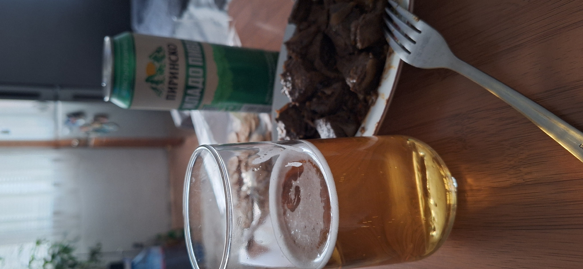 The image depicts a still life on a wooden surface. A glass of beer sits at the forefront, displaying a golden-hued liquid with a frothy head. Behind the glass is a can of "Пиринско" beer, a Bulgarian beer. Next to the beer can, on a plate, there is a portion of what appears to be meat. A fork is placed on the plate next to the meat. The background is blurred.
Given that "Пиринско" beer is a Bulgarian beer, and Blagoevgrad is a Bulgarian city, it is conceivable that this image was taken in or near Blagoevgrad. However, there is no conclusive evidence in the image to specifically place it there.