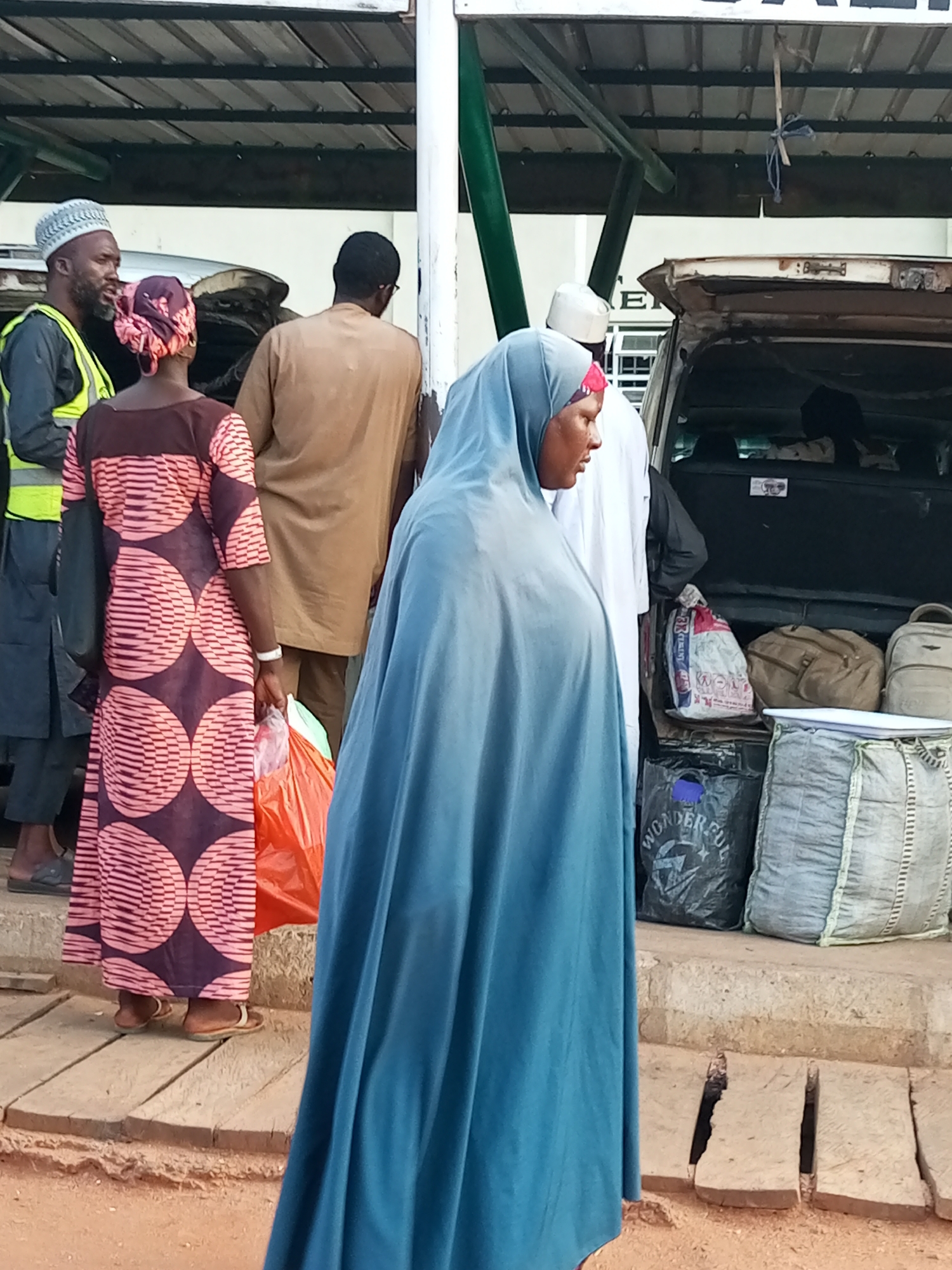 The image depicts a scene at the Gombe Central Motor Park in Gombe State, Nigeria, focusing on several individuals and their luggage. In the foreground, a woman wearing a blue hijab and a light blue abaya stands facing left, her right side partially visible. To her left, a woman in a dark purple and pink patterned dress, also wearing a pink headscarf, holds an orange plastic bag in her right hand. Behind her, a man in a tan kaftan and a white skullcap is seen from the back. To the far left, a man wearing a grey and white patterned skullcap, a grey jacket, and a bright yellow safety vest stands facing left. Behind the group, the open trunk of a vehicle reveals several bags, including a large grey woven sack, a black plastic bag with "WONDERFUL" printed on it, and two tan duffel bags. The environment suggests an outdoor transport hub with a covered waiting area overhead and a dusty, reddish-brown ground.