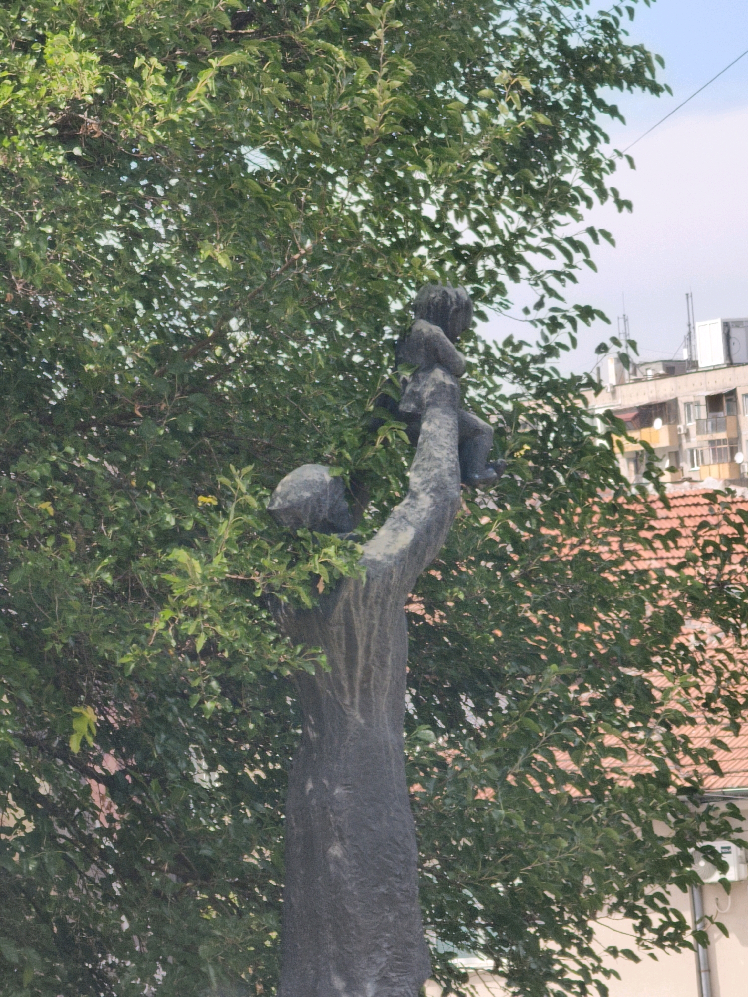 The image shows a bronze statue of a mother holding a child, positioned on a tree trunk. The statue is located in the central part of Lovech, Bulgaria, in front of the Lovech National Museum of Architecture and Construction. The statue is surrounded by lush green foliage from the tree and in the background, there are buildings with tiled roofs and air conditioning units visible. The sky is a pale blue with a few wispy clouds. The statue's rough texture and dark bronze color contrast with the vibrant green of the leaves. The mother's pose suggests protectiveness and tenderness towards the child she cradles.
