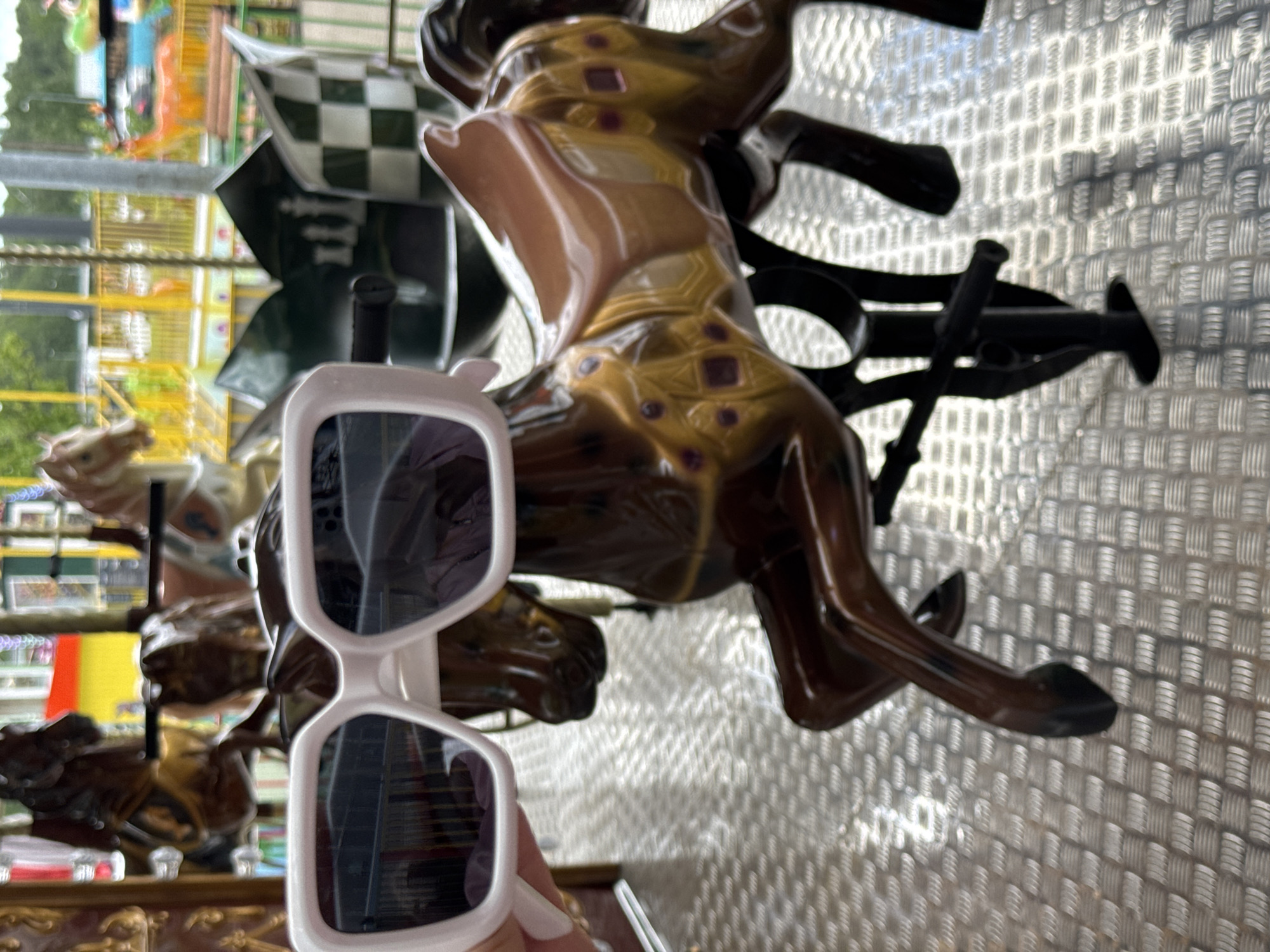 The image depicts a carousel horse with white sunglasses propped against it. The carousel appears to be in a fair or amusement park setting, evident from the blurred background showing other rides and attractions. The carousel horse is painted in brown and gold, with ornate designs. The sunglasses have a white frame and dark lenses, reflecting the surrounding environment. The background consists of metal sheeting. There is no explicit evidence to confirm the location as Saint Petersburg, Russia from the image alone.