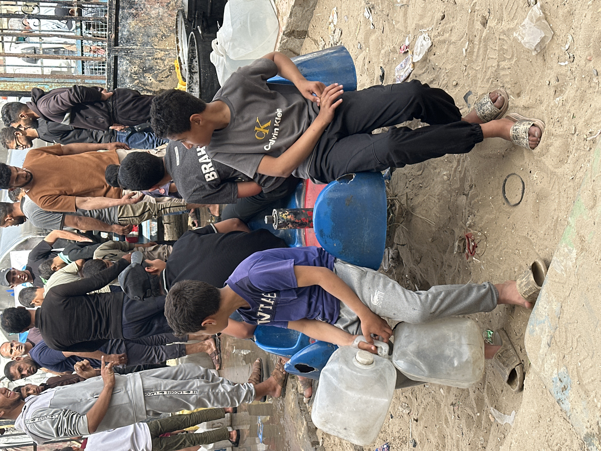 In an outdoor setting, likely Gaza, Palestinian Territories, a group of young males is gathered. Some are seated on blue plastic barrels, while others are standing or kneeling on the sandy ground. A young boy wearing a purple shirt sits on a barrel, filling plastic water jugs. Another boy in a grey Calvin Klein shirt sits nearby. The environment is littered with trash and debris. A metal fence is visible in the background. The overall impression is one of scarcity or resourcefulness.