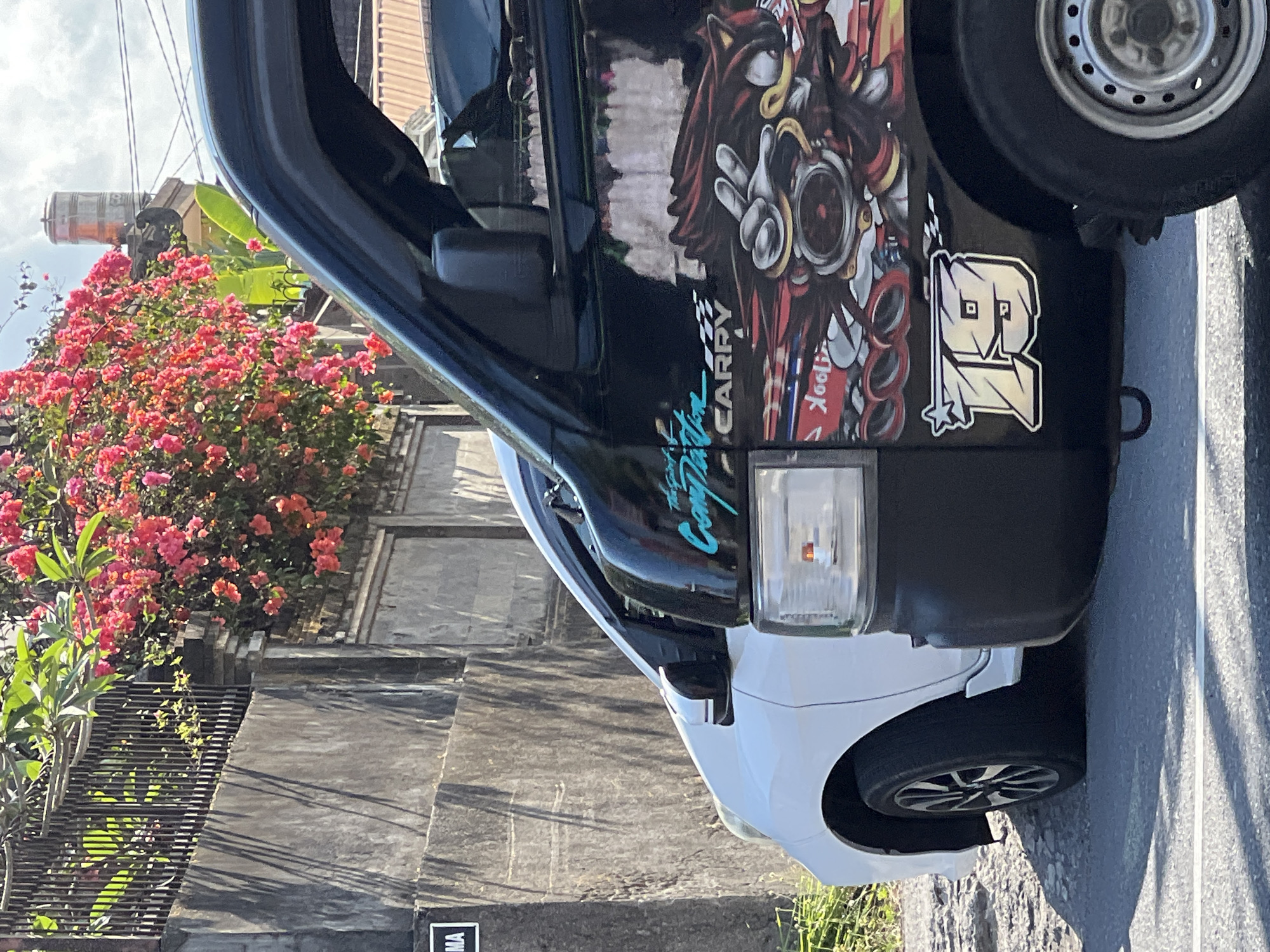 A black utility vehicle with a "CARRY" logo and a cartoon character sticker on its side is parked on an asphalt road in Denpasar, Indonesia. A white car is parked next to it. A stone wall and a fence topped with a bougainvillea bush with vibrant pink flowers are in the background. Sunlight casts shadows on the wall. A water tower and utility poles are visible in the distance.