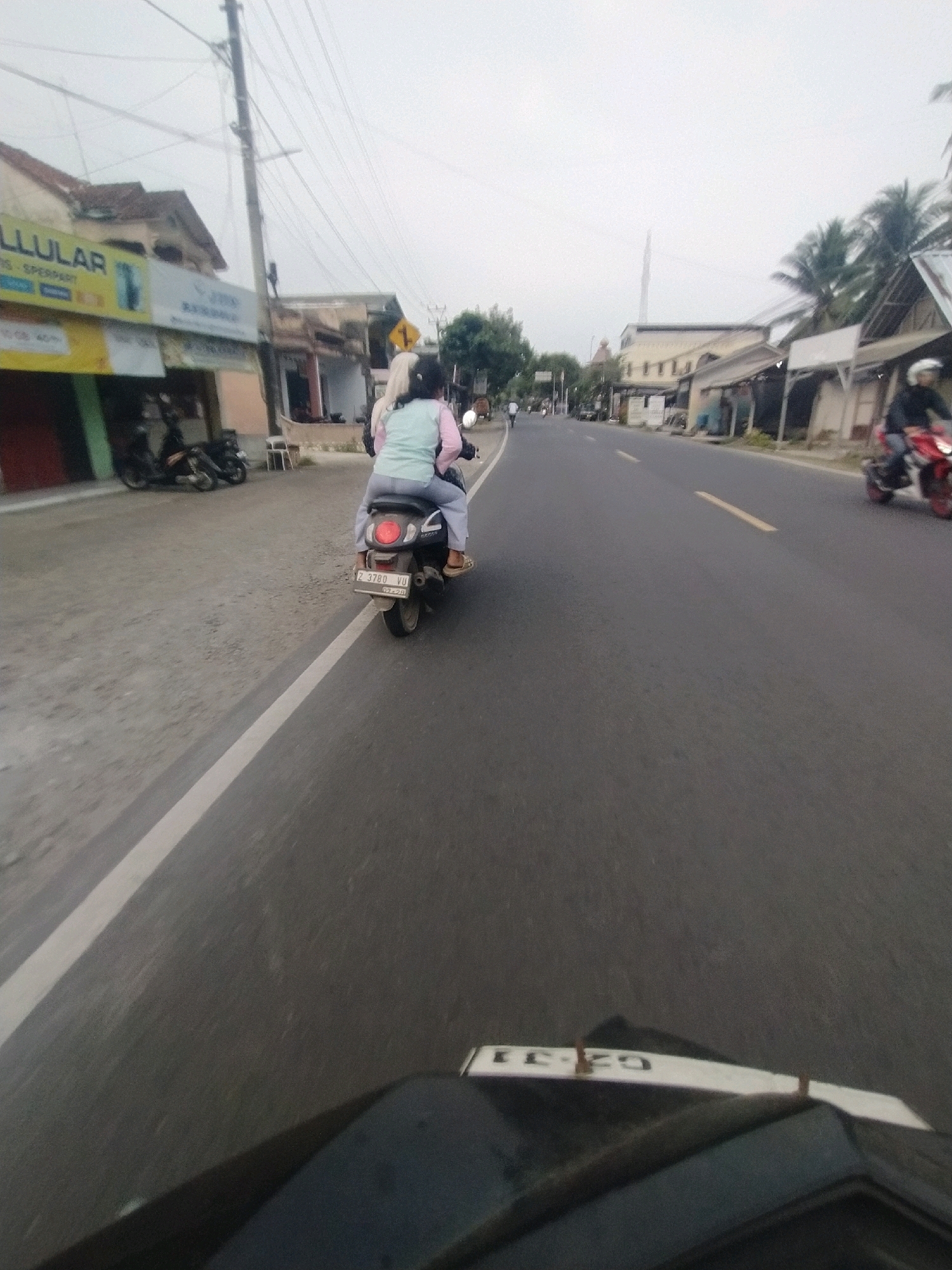 The image depicts a street scene in Desa Karangbenda, Indonesia, under an overcast sky. The perspective is from the seat of a motorcycle, looking forward and slightly to the right.
In the foreground, the black handlebar and a portion of the dashboard of the motorcycle are visible, with a white tag attached to the dashboard displaying the characters "CZ-31".
The road is paved asphalt with a white line marking the edge of the lane on the left and a yellow dashed line in the center. Traffic appears to be light.
On the left side of the road, there are several buildings with shops. One building has signage that includes the word "LLULAR" and "SPERPARY". Several parked motorcycles are visible in front of these shops. A utility pole with numerous wires stands near the sidewalk.
In the middle ground, a motorcycle with two occupants is traveling away from the camera. The person in the rear is wearing a light blue top and grey pants, with a light-colored headscarf. The person in front is wearing a pink top and appears to be wearing a helmet. The motorcycle has a license plate that reads "Z 3780 VU".
Further down the road on the right side, another motorcycle with a rider wearing a white helmet and a red and black motorcycle is visible, also traveling in the same direction. Beyond these are more buildings, some with awnings and simple structures, and a few palm trees. The background is a hazy sky, suggesting a cloudy or possibly misty day.