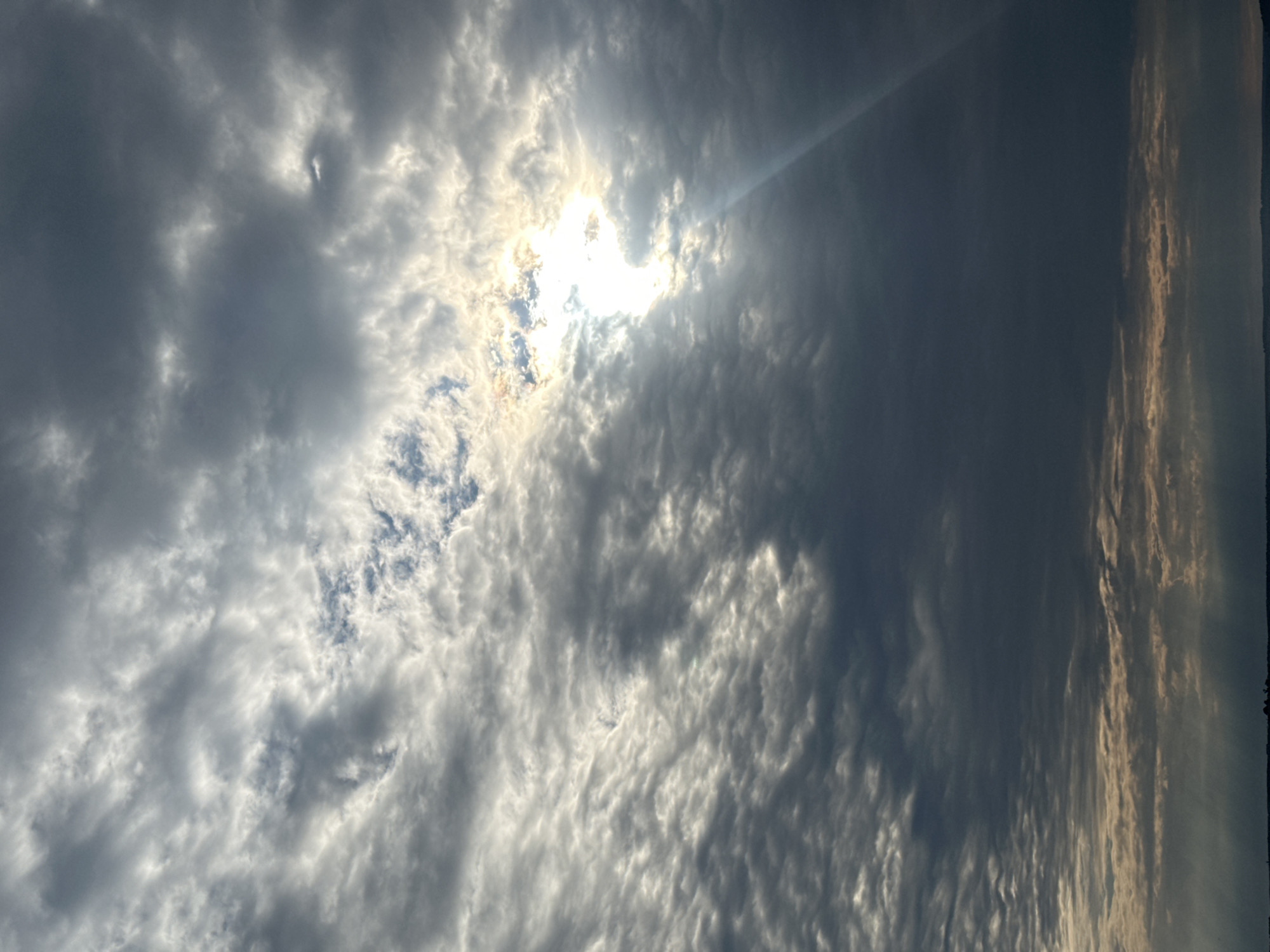 This is a photograph of the sky filled with clouds. The sun is partially obscured by the clouds, creating dramatic lighting with rays of light emanating from it. The clouds are varied in texture and density, with some appearing dark and stormy while others are bright and illuminated. The scene captures a moment of dynamic weather with light and shadow playing across the sky. There is no visible text in the image. The orientation of the photograph is vertical, with the top of the image showing a portion of the sky. The time of day appears to be daytime, given the presence of the sun. The location is Votkinsk, Russia.