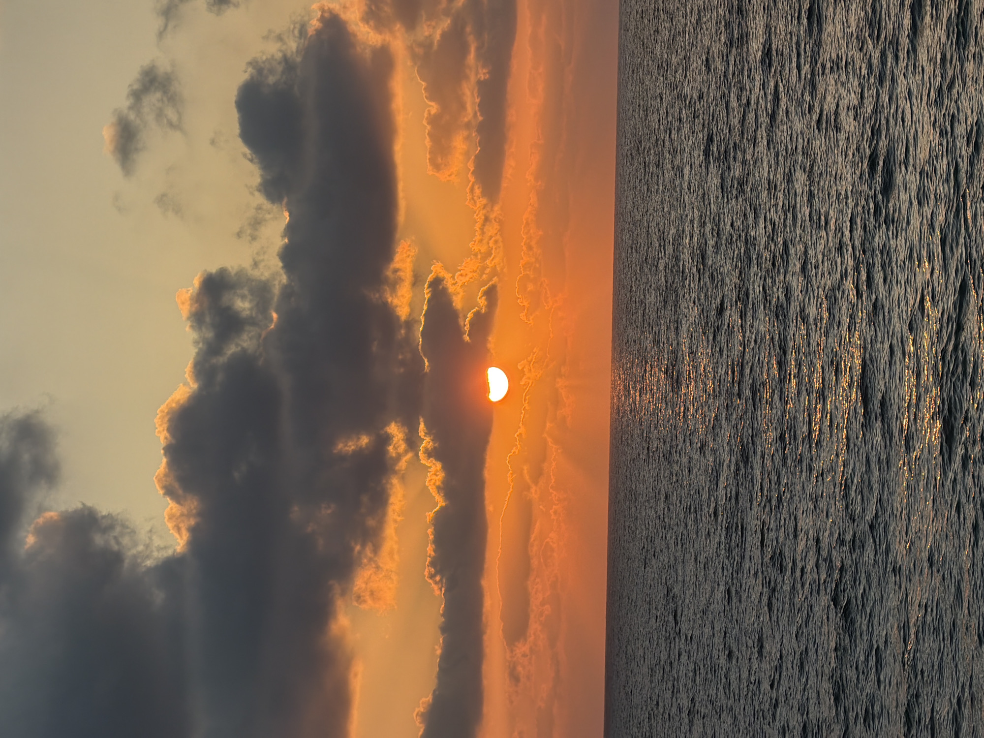 A seascape at sunset is depicted, with the sun partially obscured by clouds, casting an orange glow over the sky and water. The ocean is a deep, dark blue with ripples and reflections of the sunset. The sky is a gradient of orange and yellow, with dark gray clouds scattered across it. The image appears to have been taken from a boat or a vantage point at sea, looking towards the horizon. It is difficult to definitively ascertain the exact location in Tel Aviv without further context, but it is likely a coastal view. There are no people or specific objects identifiable in the image, making it a purely natural landscape scene.