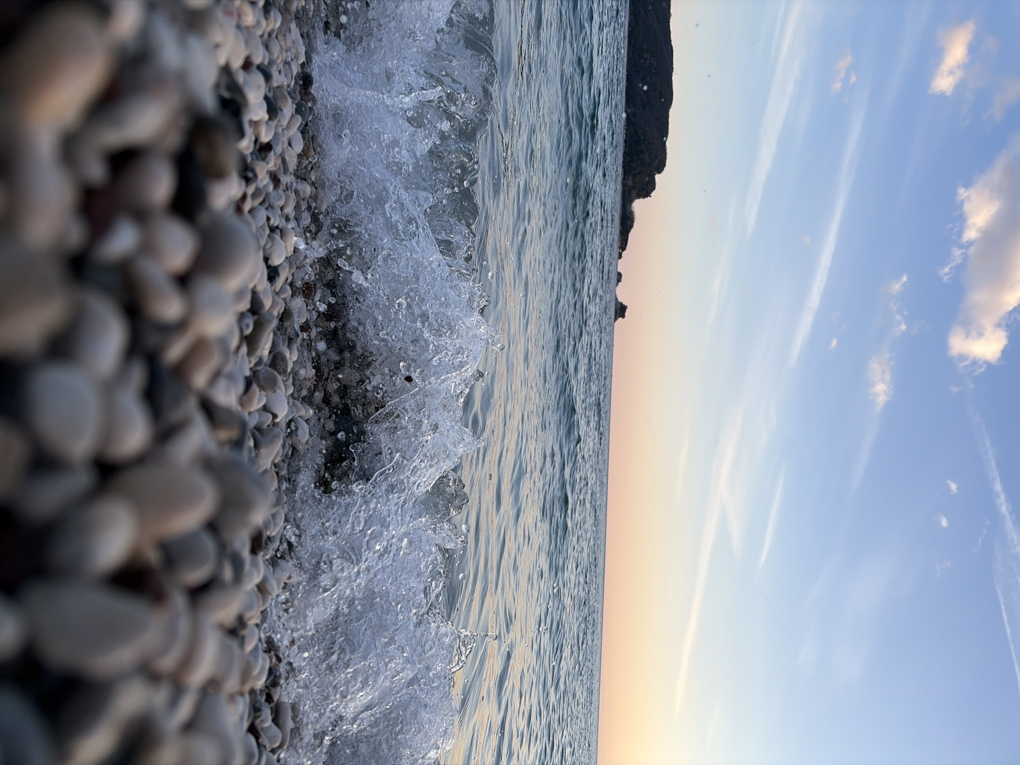 The scene depicts a low-angle view of a pebble beach in Budva, Montenegro, during twilight hours. The immediate foreground is occupied by a dense arrangement of smooth, multi-colored pebbles, ranging from off-white to dark grey and brown, with a shallow depth of field rendering the closest pebbles out of focus. A wave actively breaks across the midground, characterized by turbulent, splashing water and visible droplets. Beyond the breaking wave, the sea surface is calm with subtle ripples, displaying a turquoise-blue hue. The horizon features a large, dark, vegetated landmass, consistent with an island or peninsula, on the left, and smaller, rugged rock formations further to the right. The sky transitions from light blue overhead to a soft peach-orange at the horizon, with sparse white cirrus clouds visible. No individuals, animals, or artificial structures are present in the frame.