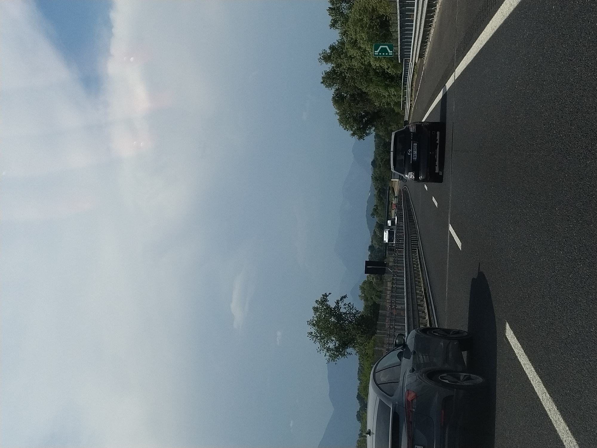 The image captures a view from a vehicle traveling on an Italian highway, likely near Felettano, Italy, given the context. The road is asphalt with clear white lane markings. In the foreground, a dark gray SUV is visible from the rear quarter, positioned in the left lane. Further ahead in the right lane, a black SUV with license plate "5XG 3233" is driving. In the distance, a truck and other vehicles are also on the highway. The landscape includes a line of trees and hills or mountains in the background. The sky is overcast with some patches of blue, and there are lens flares or light artifacts visible in the upper portion of the image. A green directional sign indicating a curve to the right is present on the right side of the road.