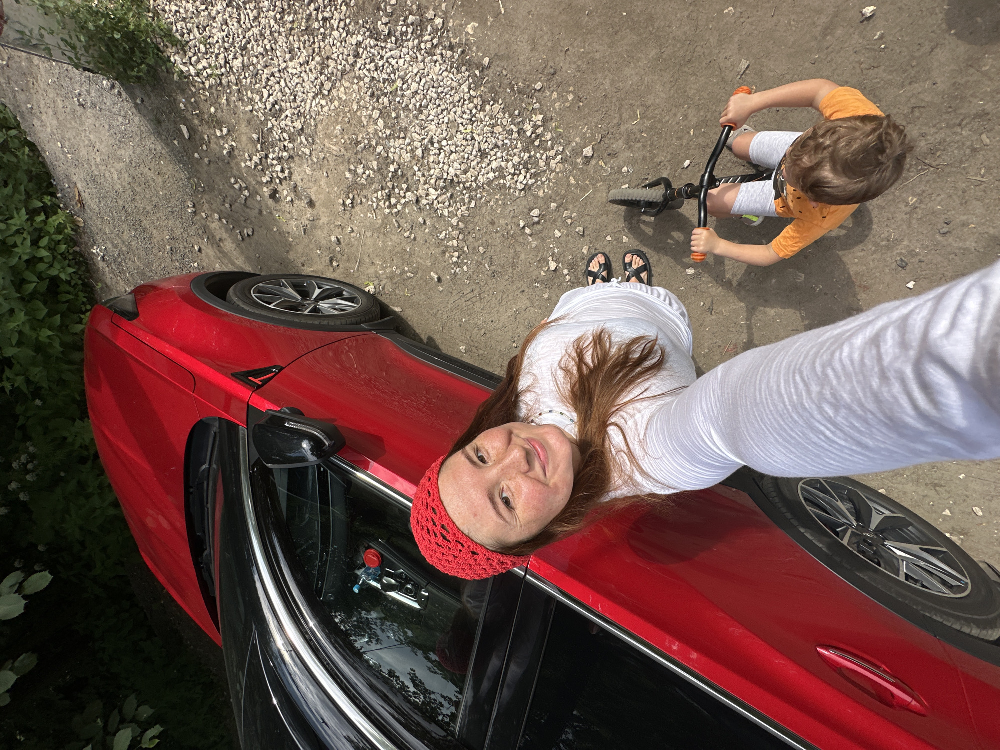 In Nizhny Novgorod, Russia, a woman in a red crocheted hat and white top takes a selfie beside a red car. Her arm is extended, holding the camera. A young child, wearing an orange t-shirt and gray shorts, rides a balance bike in the foreground, facing away from the camera. The scene unfolds on a dirt and gravel surface, with vegetation visible in the background and to the left. The sun casts shadows on the ground.