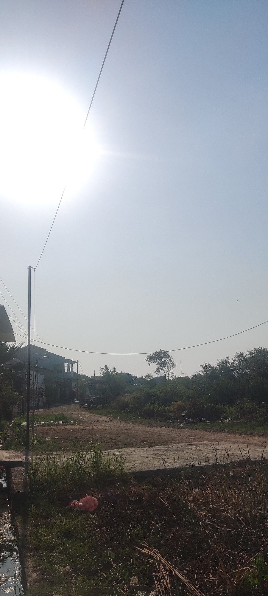 The image depicts an outdoor scene with a bright, hazy sun in the upper left quadrant. A power line stretches diagonally across the sky. In the mid-ground, a dirt road winds through a sparsely vegetated area. To the left of the road, there are a few low-rise buildings, some appearing residential or commercial, with corrugated metal roofs. A wooden structure with a thatched roof is also visible. Beyond the road, a dense line of trees and bushes forms the background, with a solitary tree standing out with a distinctive shape. The ground in the foreground is covered with dry grass and some scattered debris, including a pink plastic bag. A small, dark body of water is present on the far left edge of the image, bordered by a concrete edge. The scene appears to be in a rural or semi-urban area, consistent with a location in Gebang, Indonesia, characterized by a dusty road, modest structures, and abundant vegetation under a bright, possibly humid, sky. There are no people visible in the image.