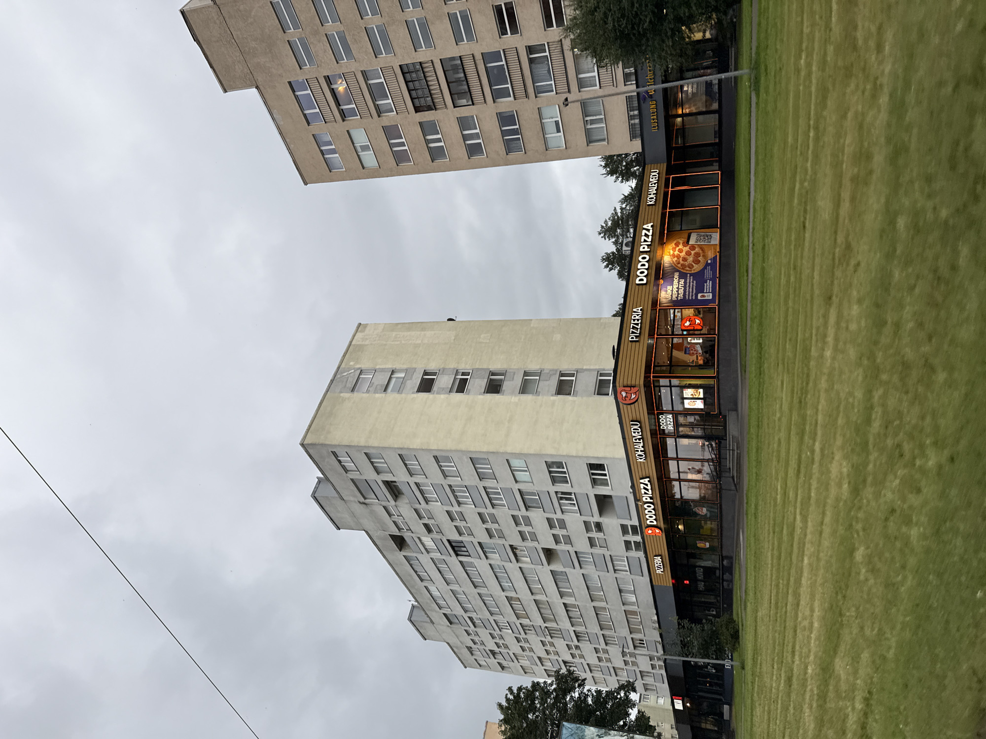 The image shows a street-level view of a commercial area in Tallinn, Estonia, with two large apartment buildings dominating the background. The foreground features a well-maintained grassy lawn. In the middle ground, a row of businesses is visible, most notably a "DODO PIZZA" restaurant with illuminated signage and storefront windows displaying pizzas and advertisements. Adjacent to the pizza place are other establishments, including a beauty salon ("ILUSALONG"). The apartment buildings are characteristic of Soviet-era architecture, one taller and lighter colored, the other slightly shorter and darker. The sky is overcast, suggesting a cloudy day. No people are clearly visible in the image.