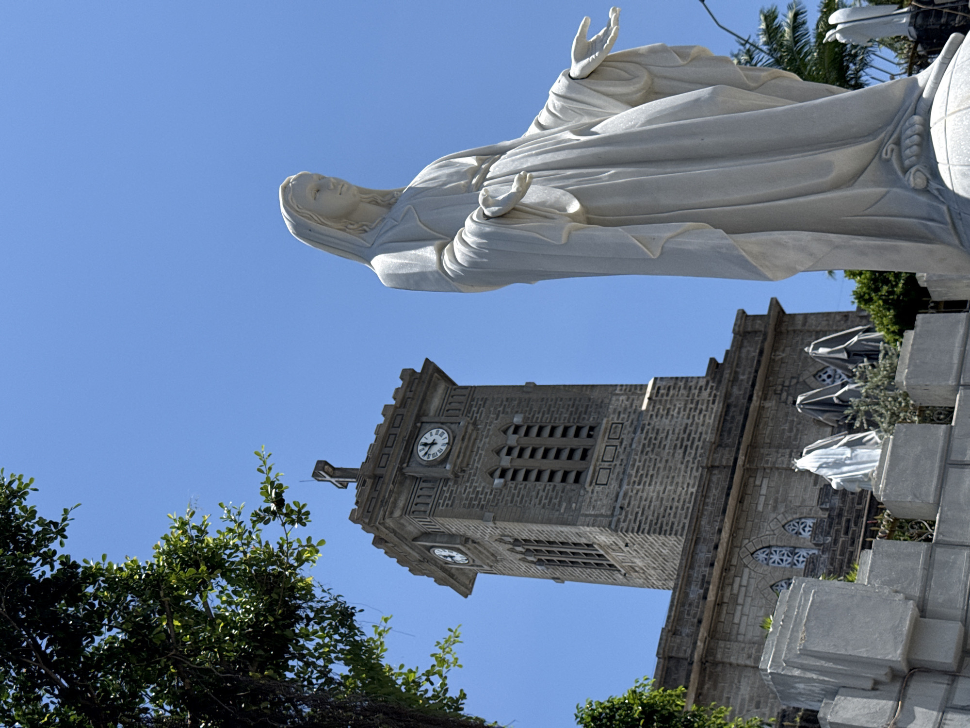 The image depicts a stone statue of the Virgin Mary, rendered in white marble, positioned in the foreground and angled to the right. The statue is life-sized and stands with its arms outstretched. Behind the statue, a stone church tower with a clock face and a cross on top is visible. The tower is constructed of gray stone blocks and features several windows. The church facade, also made of stone, has arched windows. A lush green tree branch extends from the upper left corner of the frame, partially obscuring the sky. The sky is a clear, bright blue. The scene is set outdoors, under daylight conditions, with the sun casting shadows. The context provided indicates this scene is located in Nha Trang Ward, Vietnam.