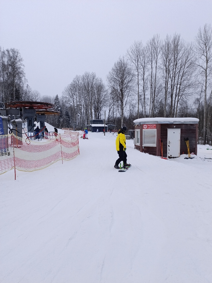 The image captures a snowy winter scene at a ski resort under an overcast sky. The ground is covered in white snow, showing various tracks and patterns from skiing and snowboarding activities.
In the immediate foreground and midground, a person wearing a bright yellow jacket and black pants is standing on a snowboard, facing slightly to the right and away from the camera. The snowboard is green and black. To their left, a red and white safety net or fence stretches across the snow, acting as a boundary.
To the right of the snowboarder, a small, rustic wooden building with a rounded roof is visible. The building is brown with white trim around its windows and a white door. A red sign with a white cross, the universal symbol for first aid, is prominently displayed above one of the windows. Below this symbol, Cyrillic text is visible, which reads "СПАСАТЕЛИ" (Rescuers) and "ПУНКТ ПОМОЩИ" (Aid Post), along with a website address "WWW.PRUDKI.RU". Leaning against the side of this building are several red shovels and yellow poles or brooms.
Further into the scene, to the left, a large ski lift mechanism with grey support structures and red operational components is visible. Several individuals are gathered near the base of this lift, some appearing to be waiting in line or preparing to use it. Another person, dressed in a blue jacket and yellow pants, is seen on skis further back in the midground, near a smaller, darker building which is partially obscured by trees.
The background is dominated by a dense forest of tall, bare deciduous trees, characteristic of a winter landscape. A few evergreen trees can also be seen interspersed among them. The sky is a uniform light grey, suggesting an overcast day. The overall impression is that of a functional, active ski area during winter.
