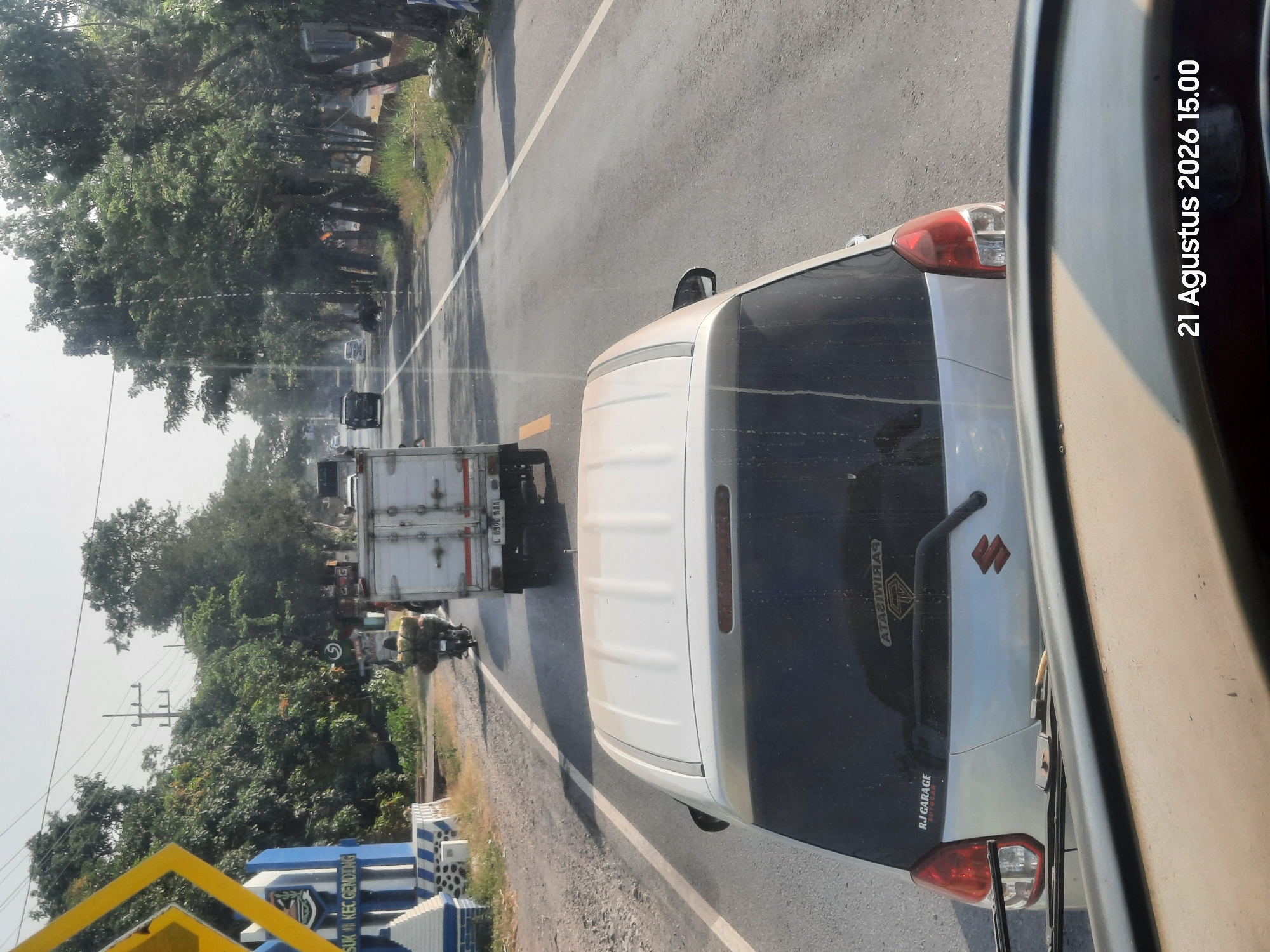 The image depicts a street scene in Karangpranti, Indonesia, captured on August 21, 2026, at 15:00. In the foreground, the rear of a light-colored car is visible, with its windshield and roofline occupying the bottom portion of the frame. The car has a visible rear spoiler and a Suzuki logo on the trunk. A sticker reading "RJ GARAGE" is present on the left side of the rear bumper.
The car is on a paved road, with a white lane divider line to its right. Traffic is moving in the same direction. Ahead of this car, a white box truck is in the adjacent lane. The truck has a license plate that appears to read "L 6590 BAA". Further ahead, a motorcycle carrying a large load of what appears to be foliage or produce is also in the lane.
To the left of the road, a blue and white striped monument or signpost is visible, with text that appears to say "SUK KEC GENDING". This monument is bordered by a low wall and some vegetation.
The environment is characterized by a road lined with numerous trees, suggesting a somewhat rural or suburban setting. The sky is clear and bright, indicating a sunny day. Power lines are visible suspended over the road. The overall scene suggests ordinary daytime traffic and activity in a roadside area of Karangpranti.