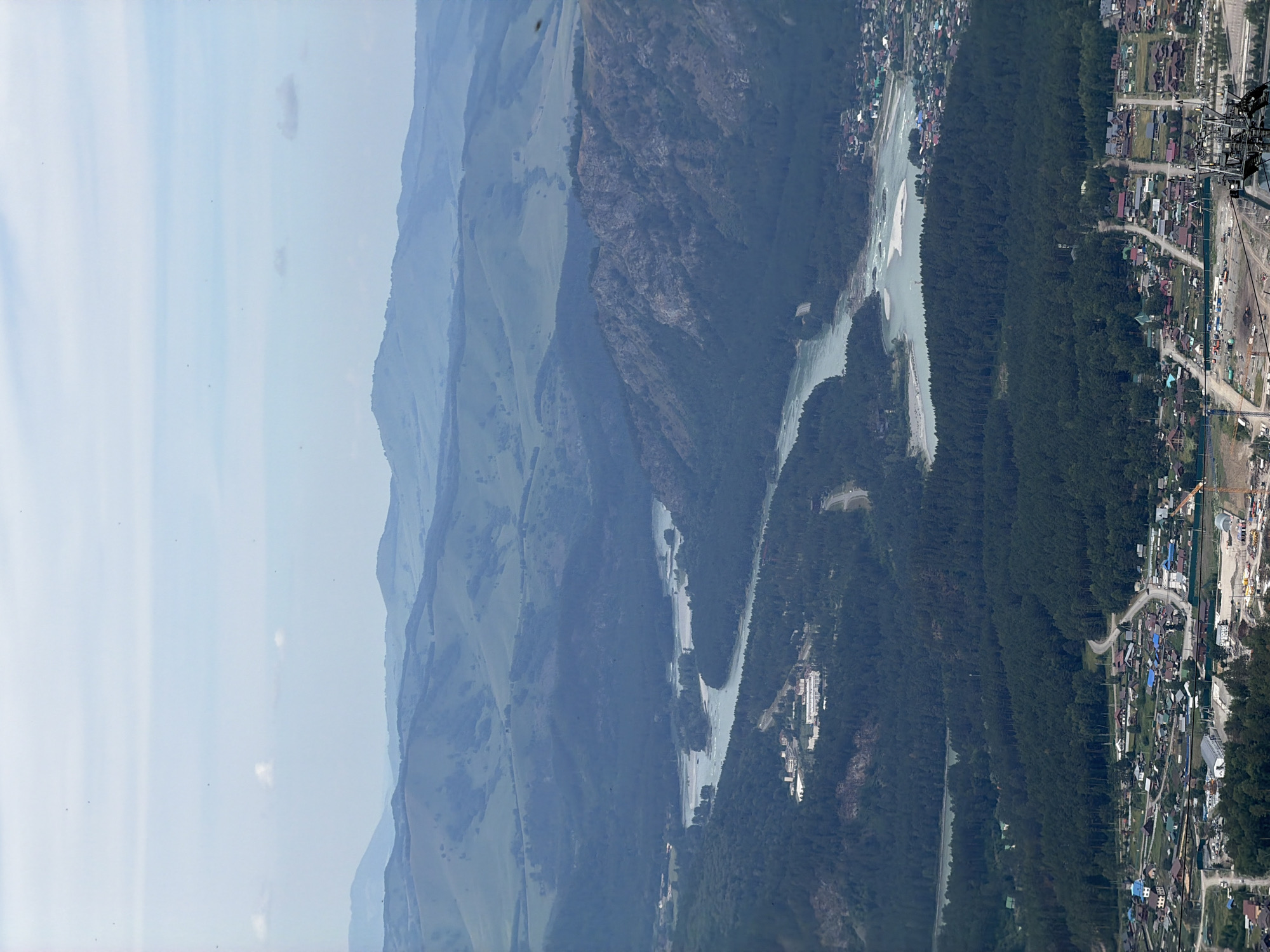 An aerial, wide-angle view captures a sprawling, verdant landscape in Ozernoe, Russia, characterized by rolling, tree-covered hills and a winding, turquoise river. The distant mountains are shrouded in a soft, blue haze, indicating their immense distance. The middle ground is dominated by a dense, dark green forest that gives way to the vibrant river, which flows through a series of islands and sandbars. Closer to the foreground, a village with numerous houses, roads, and some construction activity, including a crane, is visible. A few buildings, possibly a resort or industrial complex, are situated on the edge of the forest along the river. The sky is a pale blue with scattered white clouds. The overall scene is one of natural beauty and human settlement integrated into the environment.