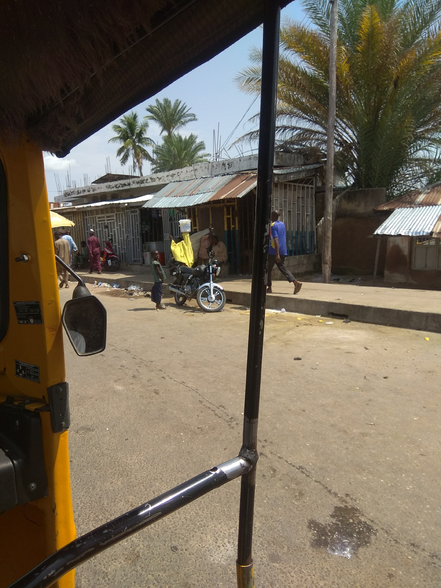 This image depicts a street scene in Jalingo, Nigeria, likely a market or commercial area. In the foreground, the side of a yellow auto-rickshaw (tricycle taxi) is visible, with its thatched roof and part of the driver's cabin. A black metal handlebar extends across the lower part of the frame.
In the middle ground, a street runs towards the background. A young boy in a green shirt and dark trousers stands on the right side of the road, facing left. A parked motorcycle with a yellow cloth draped over its handlebars is to his right. Further back, on the left side of the street, several people are walking or standing. One person in a red robe is prominent.
The background reveals a row of single-story buildings with corrugated metal roofs, typical of commercial structures in such areas. Palm trees are visible among the buildings, indicating a tropical climate. The sky is clear and blue. The road surface appears to be asphalt with some patches of dirt and debris. The overall impression is a bustling, ordinary day in a Nigerian city.