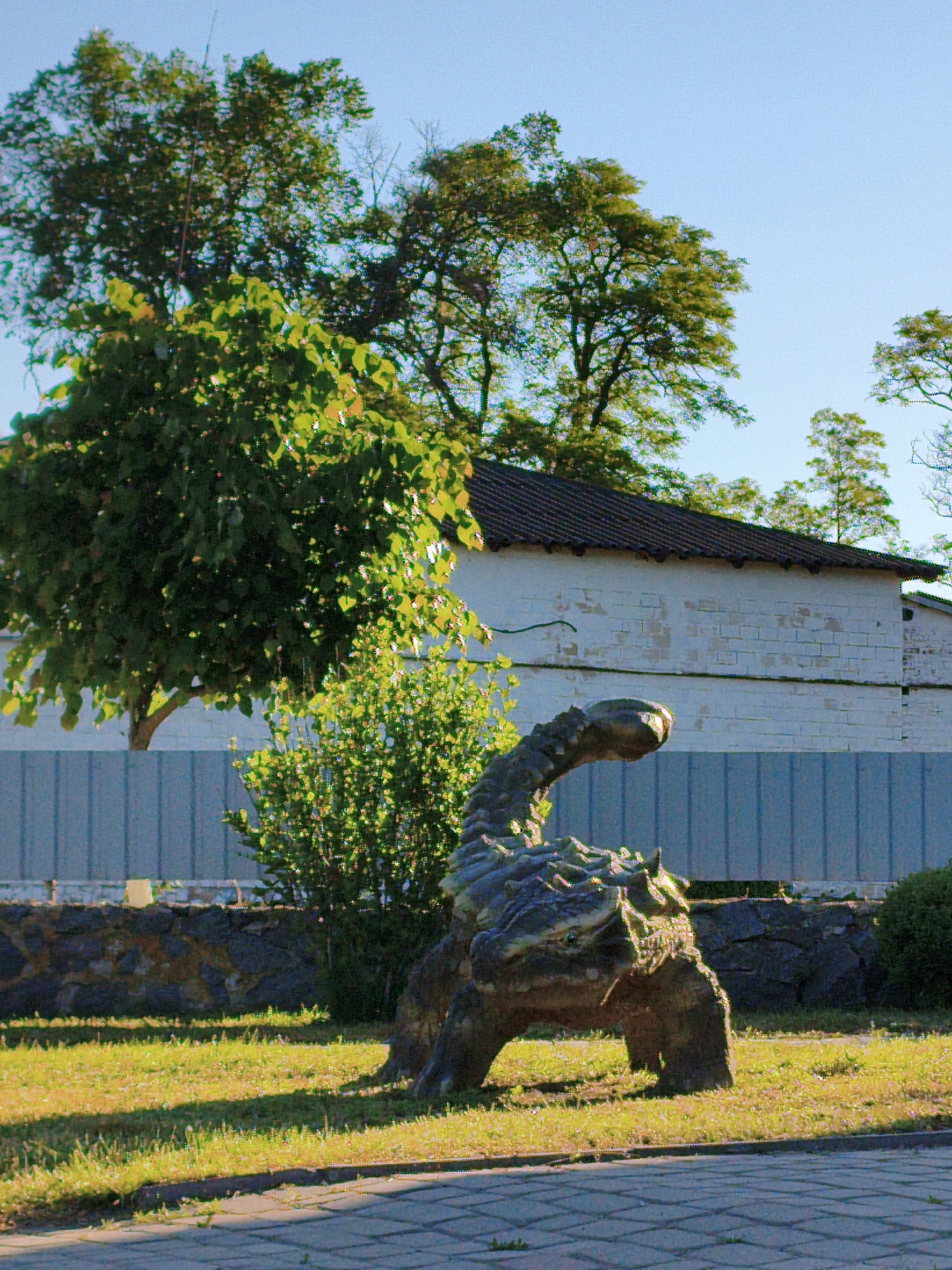 The image shows a bronze sculpture of a dinosaur, possibly an ankylosaurus, in a park-like setting. The sculpture stands on a patch of green grass adjacent to a paved pathway. Behind the sculpture, a low stone wall and a blue metal fence run along the background, partially obscured by bushes. A single-story white building with a red tile roof is visible behind the fence and trees. The sky is clear and blue. The image was captured in Ustyvytsia, Ukraine. There are no people present in the image.