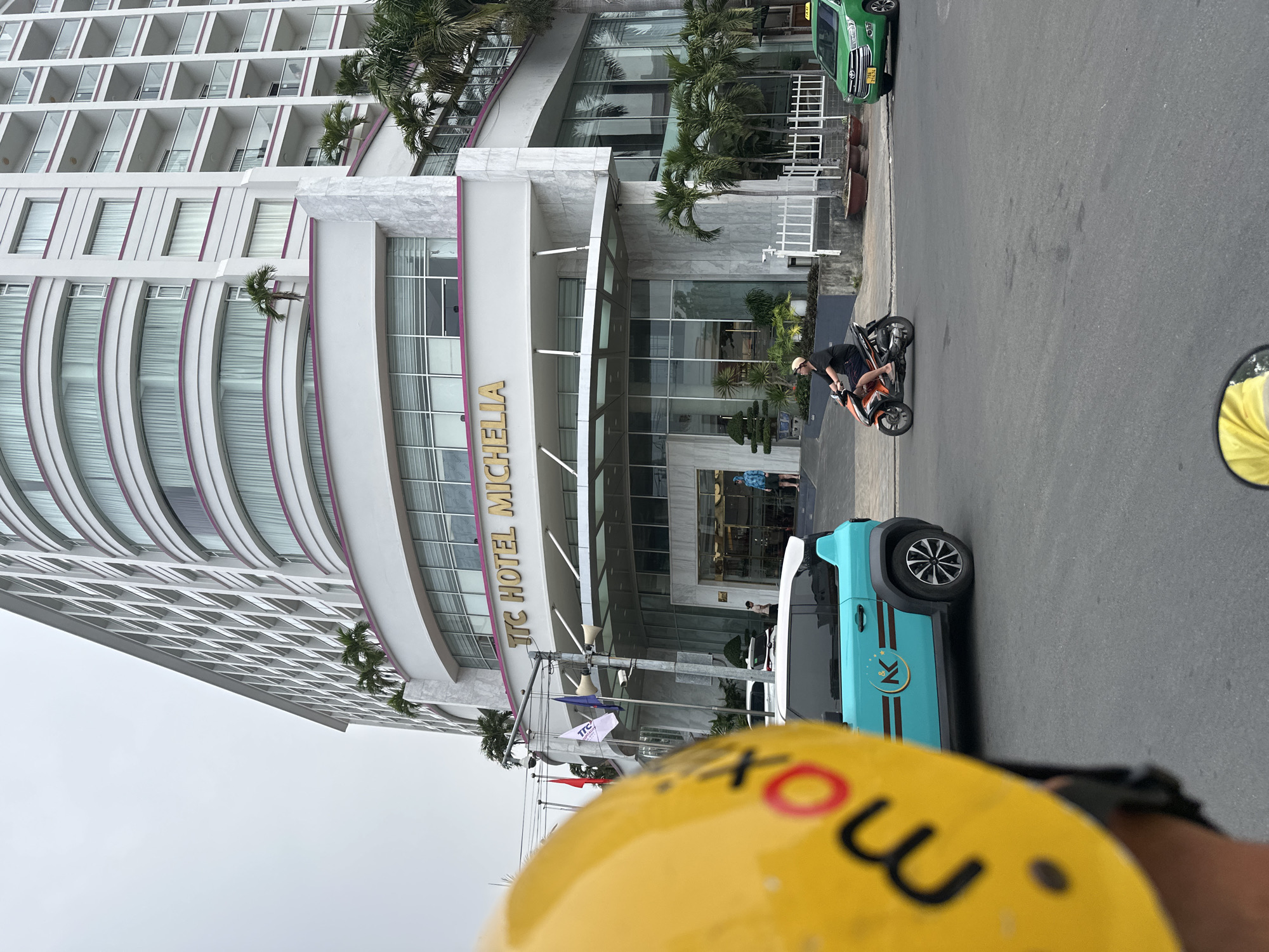 The image captures the TTC Hotel Michelia in Nha Trang Ward, Vietnam. The hotel is a tall, white building with curved tiers and purple accents. Its entrance is visible, with the hotel's name displayed prominently. In front of the hotel, on the street, a person is on a motorcycle. A light blue car is parked on the street, bearing a logo with a star and the letter "K". A green car is visible further down the street. The image is taken from a first-person perspective, as a yellow helmet with black writing is visible in the foreground.