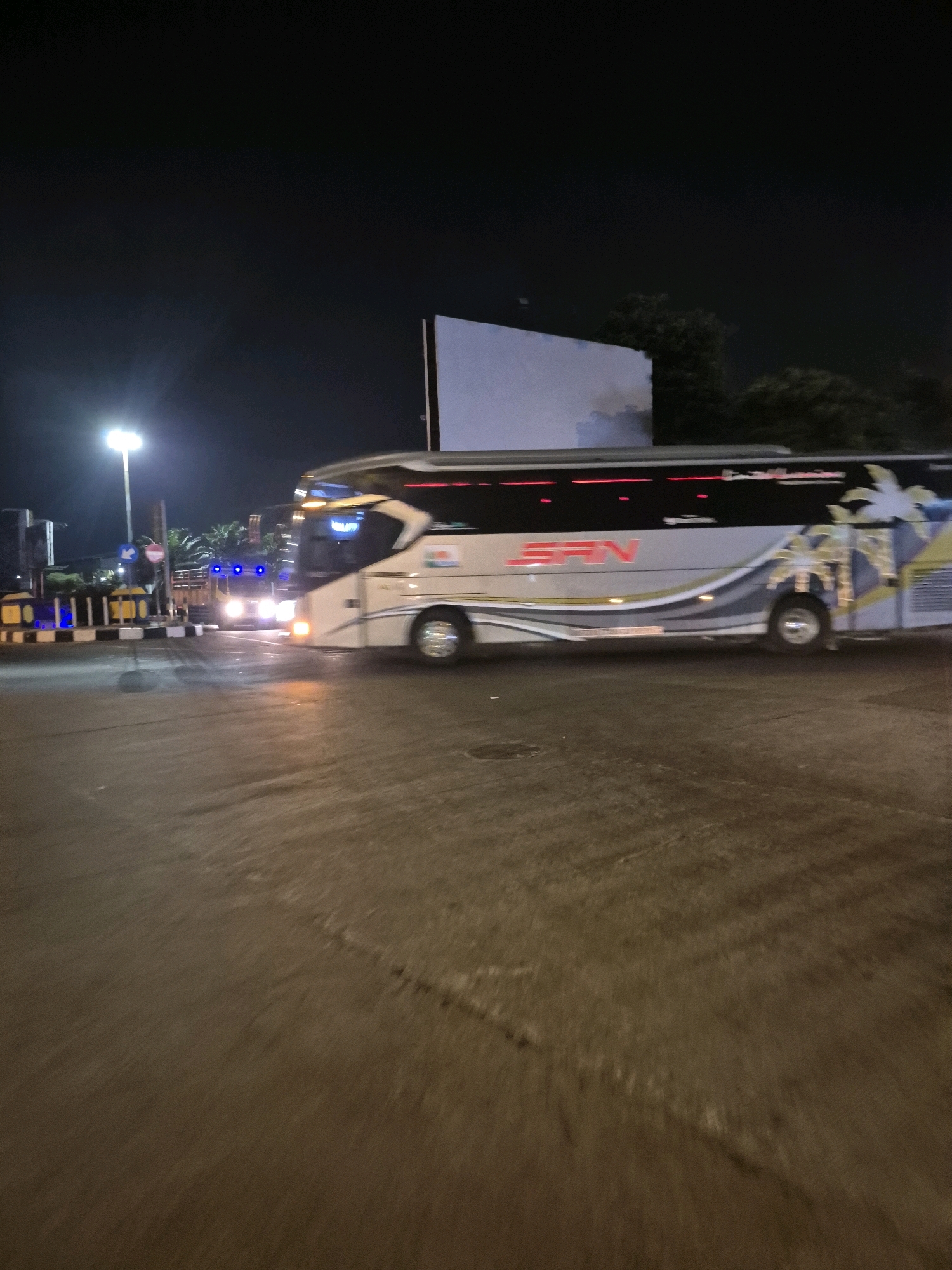 This image captures a nighttime scene at a bus terminal in Surakarta, Indonesia. A large, white bus with blue and yellow accents is parked, its headlights illuminating the wet pavement. The bus features text in a foreign script and palm tree graphics along its side. To the left, a smaller truck with bright blue lights is visible. The background includes a tall street lamp, palm trees, and a large white billboard. The overall impression is one of a typical transportation hub operating in the evening hours.