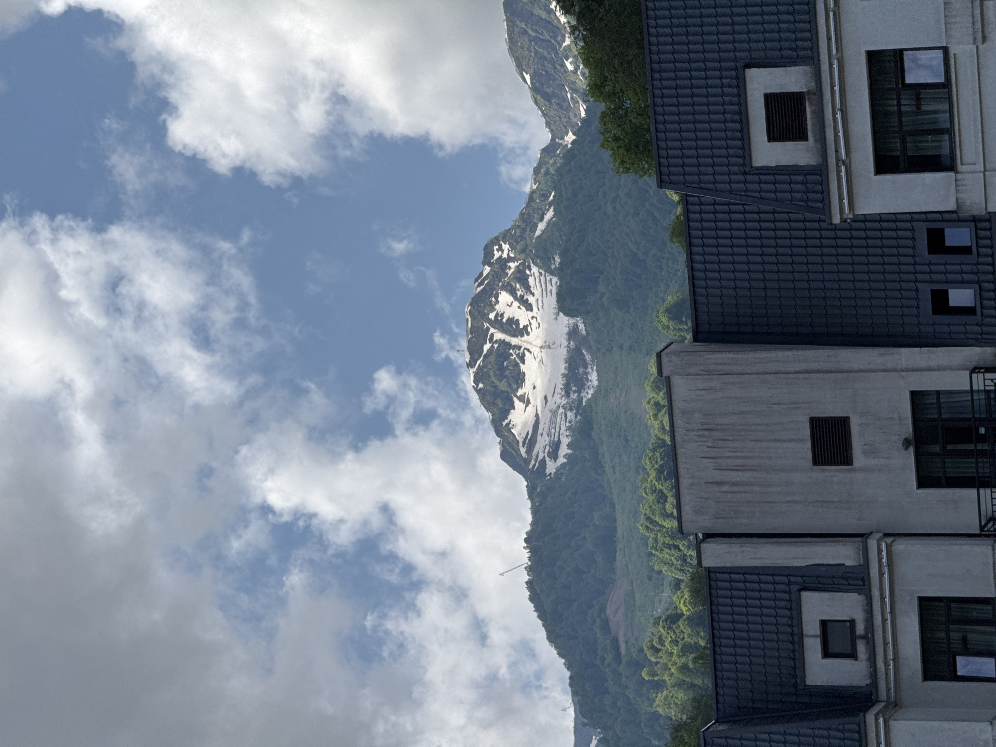 The image shows a view of a snow-capped mountain with green trees in the foreground, and buildings with dark tiled roofs. Two cranes are visible on the mountainside. The sky is partly cloudy with blue patches. The architecture of the buildings suggests a resort or residential area. This scene is consistent with the mountain landscape of Estosadok, Russia.